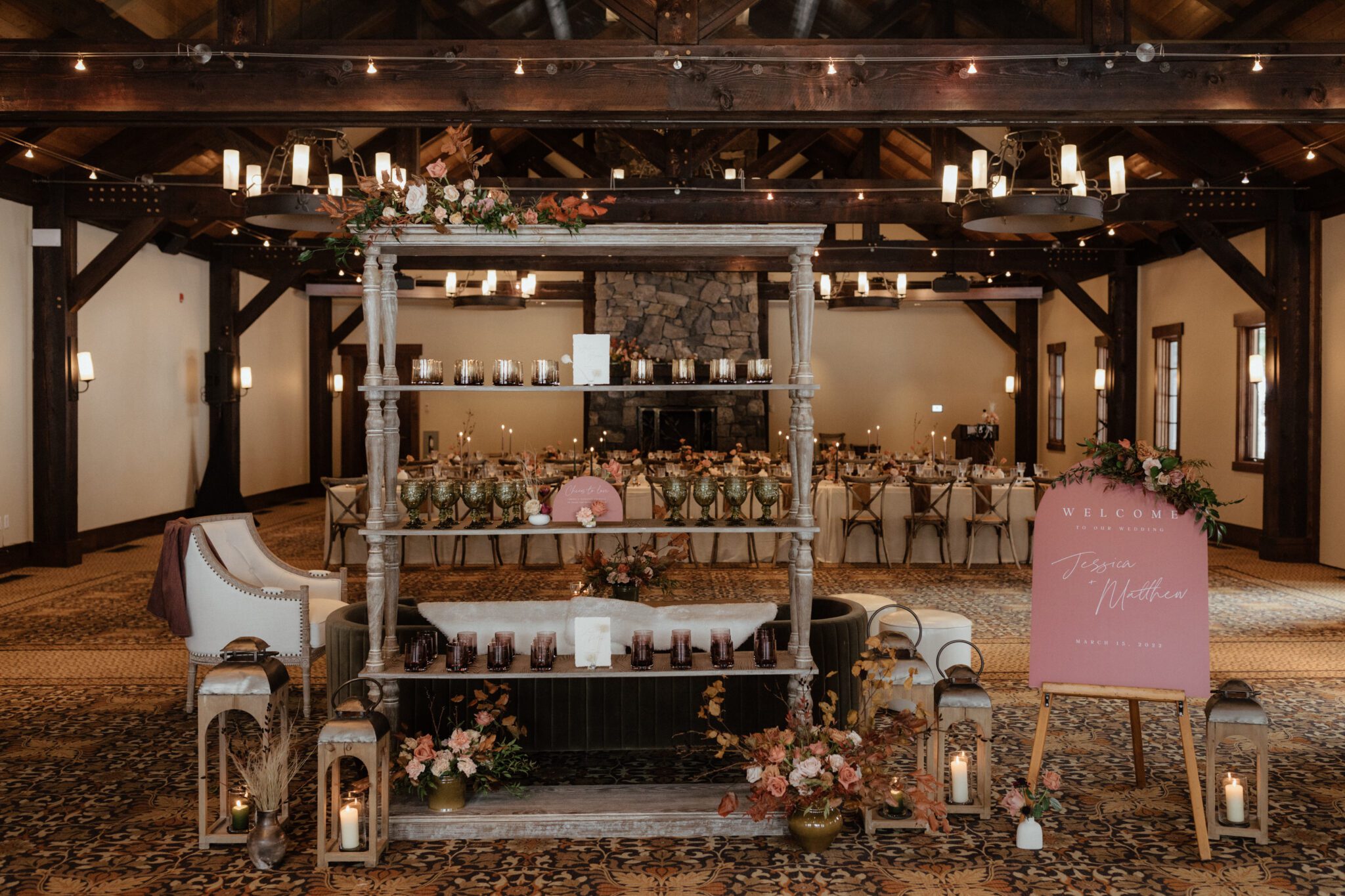 A rustic wedding reception venue in Canmore Alberta, Silvertip Resort, featuring a wooden shelf with candles and decorations. In the background, a long dining table is elegantly set. A welcome sign on an easel is to the right. Soft lighting and floral arrangements complete the cozy atmosphere.