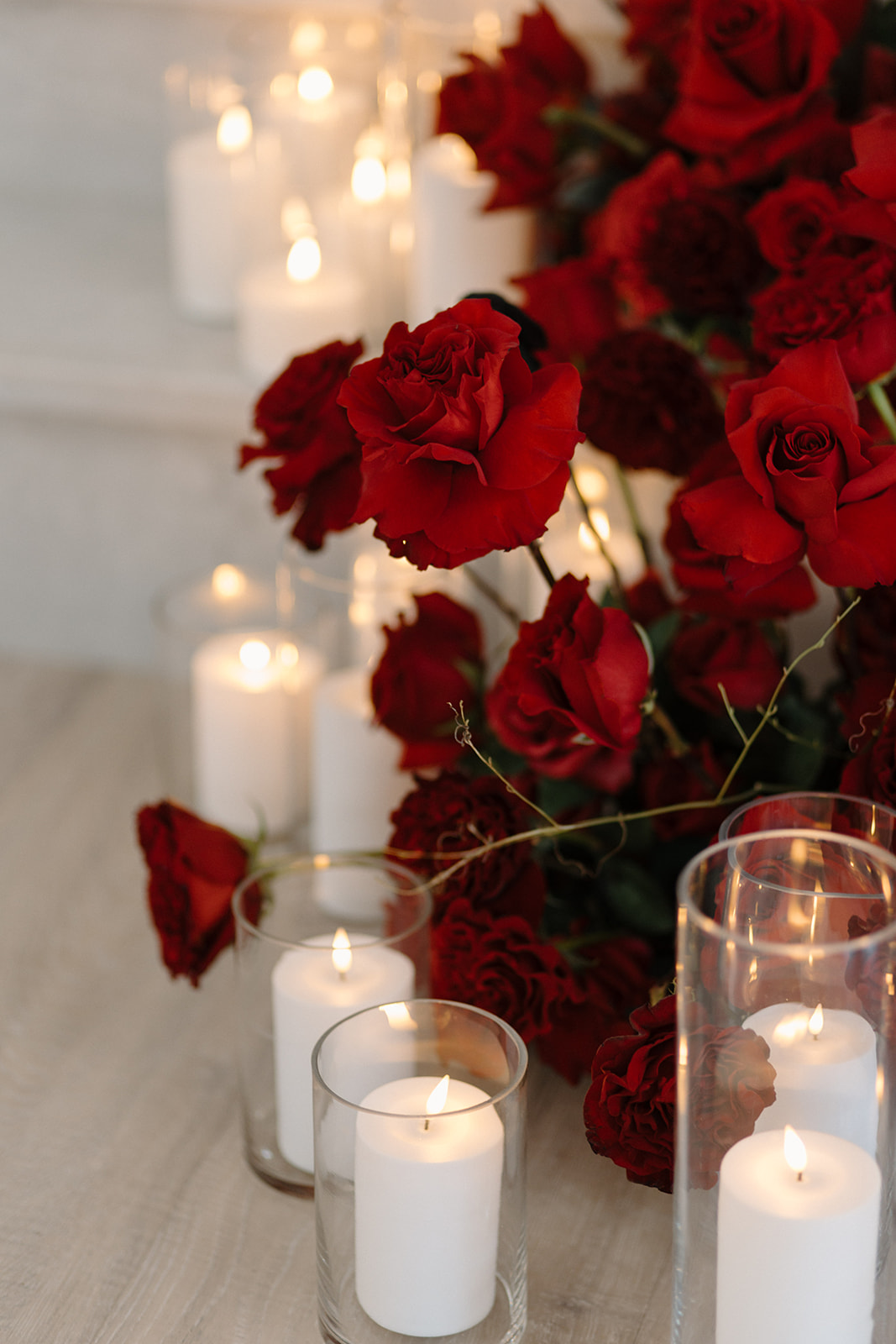 A romantic arrangement of vibrant red roses and glowing white candles in glass holders placed on a wooden surface. The soft candlelight illuminates the flowers, creating a warm and inviting ambiance.