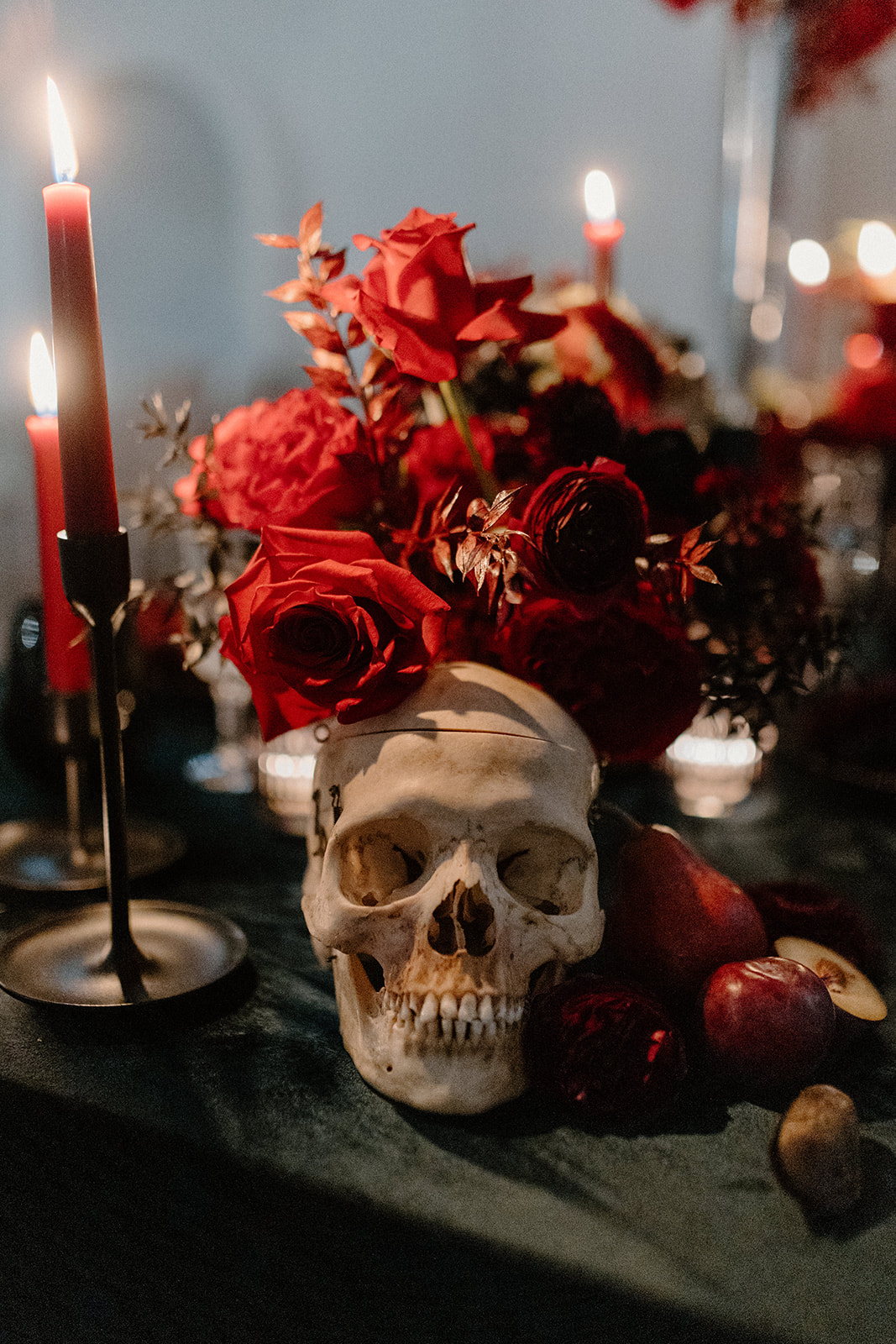 A Halloween-themed arrangement with a skull, red roses, candles, and fruit set on a dark surface, creating a spooky yet elegant atmosphere.