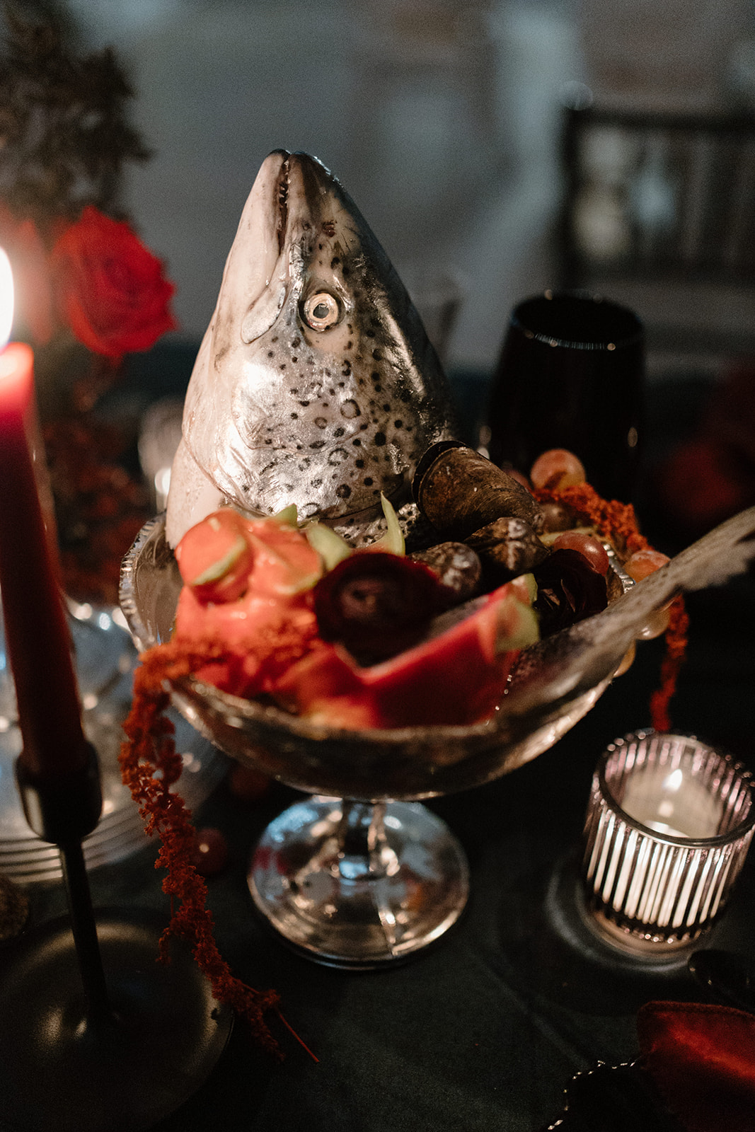 A decorative platter on a dark table displays a fish head propped upright among various fruits and vegetables. Candles are lit nearby, creating a warm ambiance.