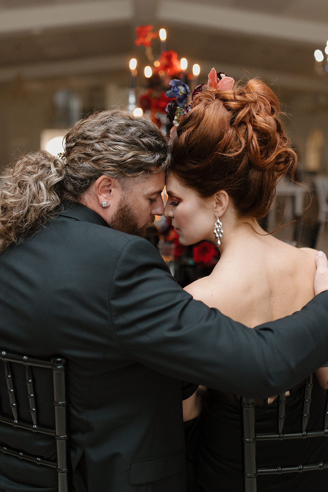 A couple in formal attire sits closely, facing each other, and touching foreheads. The woman has an elaborate updo with flowers, and both have their arms around each other. Red roses and soft lighting create a romantic ambiance.