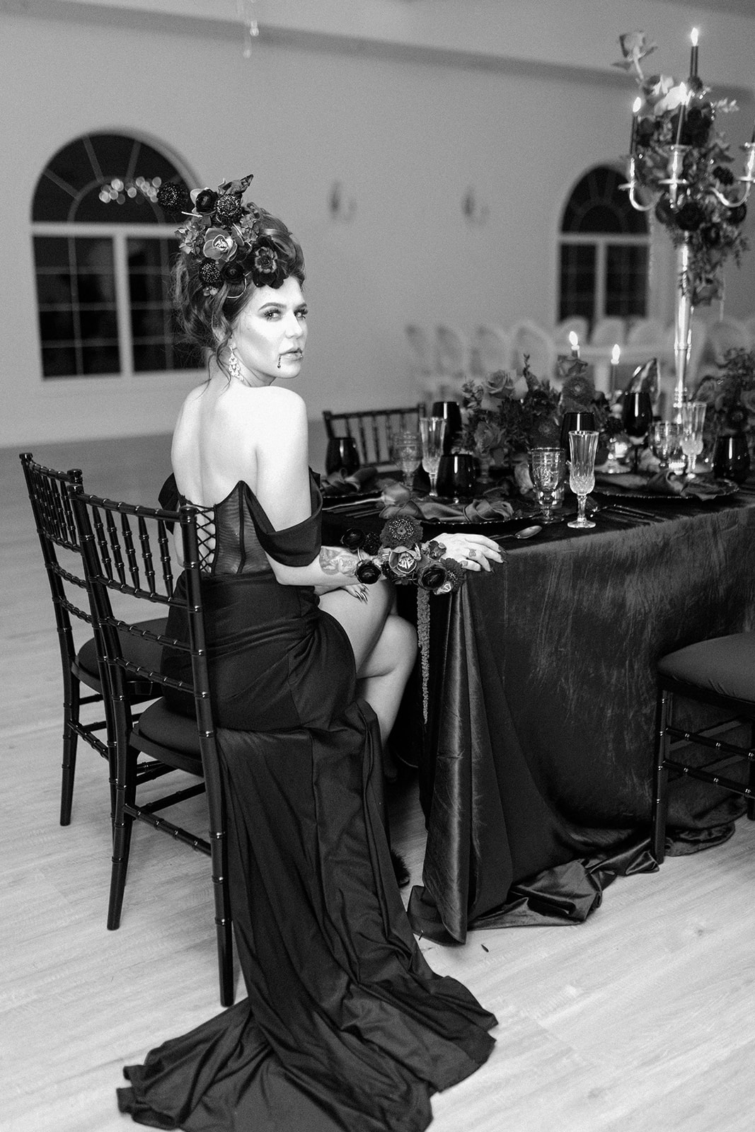 A woman in an elegant off-shoulder black dress with a long train and floral headpiece sits at a festively set dining table. The room is decorated with tall candle holders and dark table linens, creating a sophisticated atmosphere.