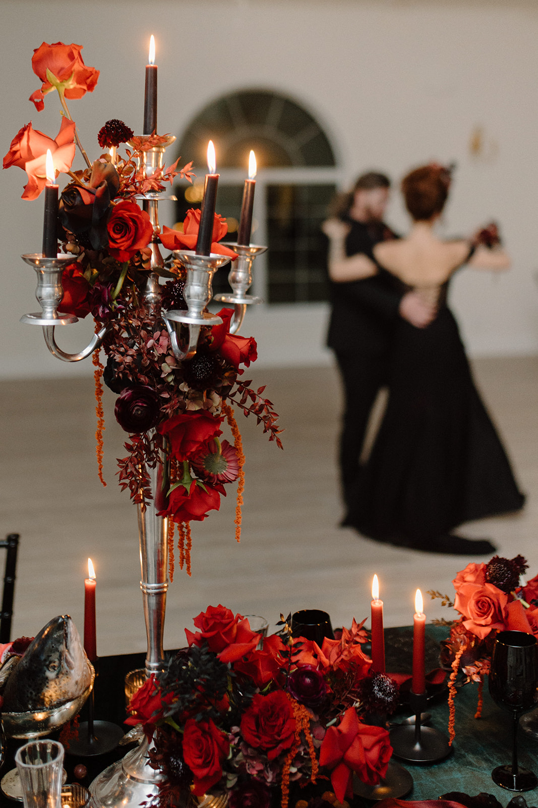 A dimly lit ballroom features a table adorned with red and orange roses, candles, and a silver candelabrum. In the blurred background, two people in formal attire dance gracefully.