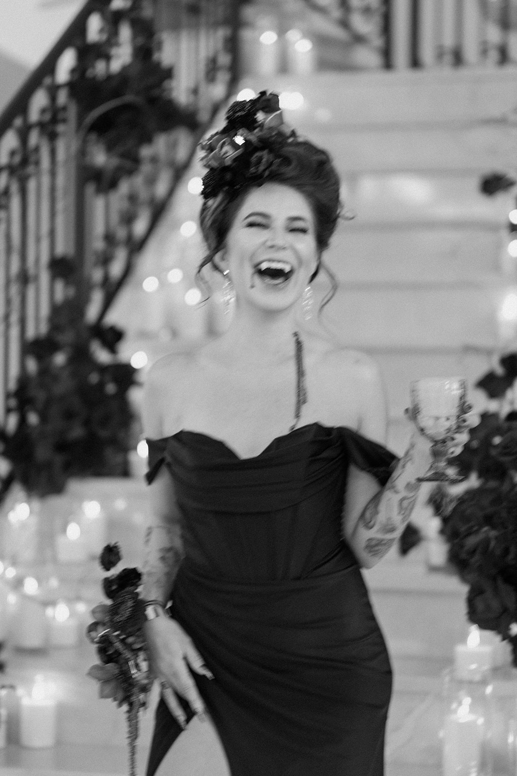 A woman in a dark dress laughs while holding a drink in one hand and flowers in the other. She stands in front of a staircase adorned with candles and flowers. The image is in black and white. A Bite of Dark Romance & Gothic Glamour in This Dracula Dinner Party Inspiration