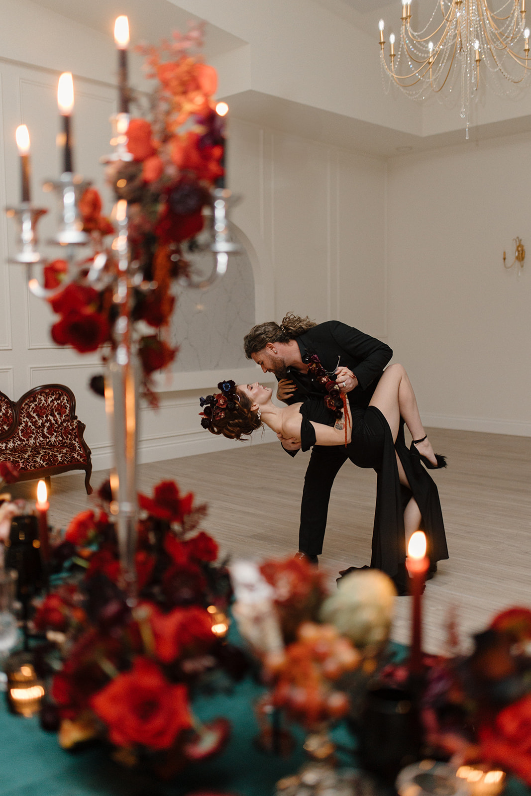 A couple dances passionately in an elegantly decorated room. The woman wears a black dress with a floral headpiece, while the man is dressed in black. The foreground features lush red floral arrangements and lit candles.