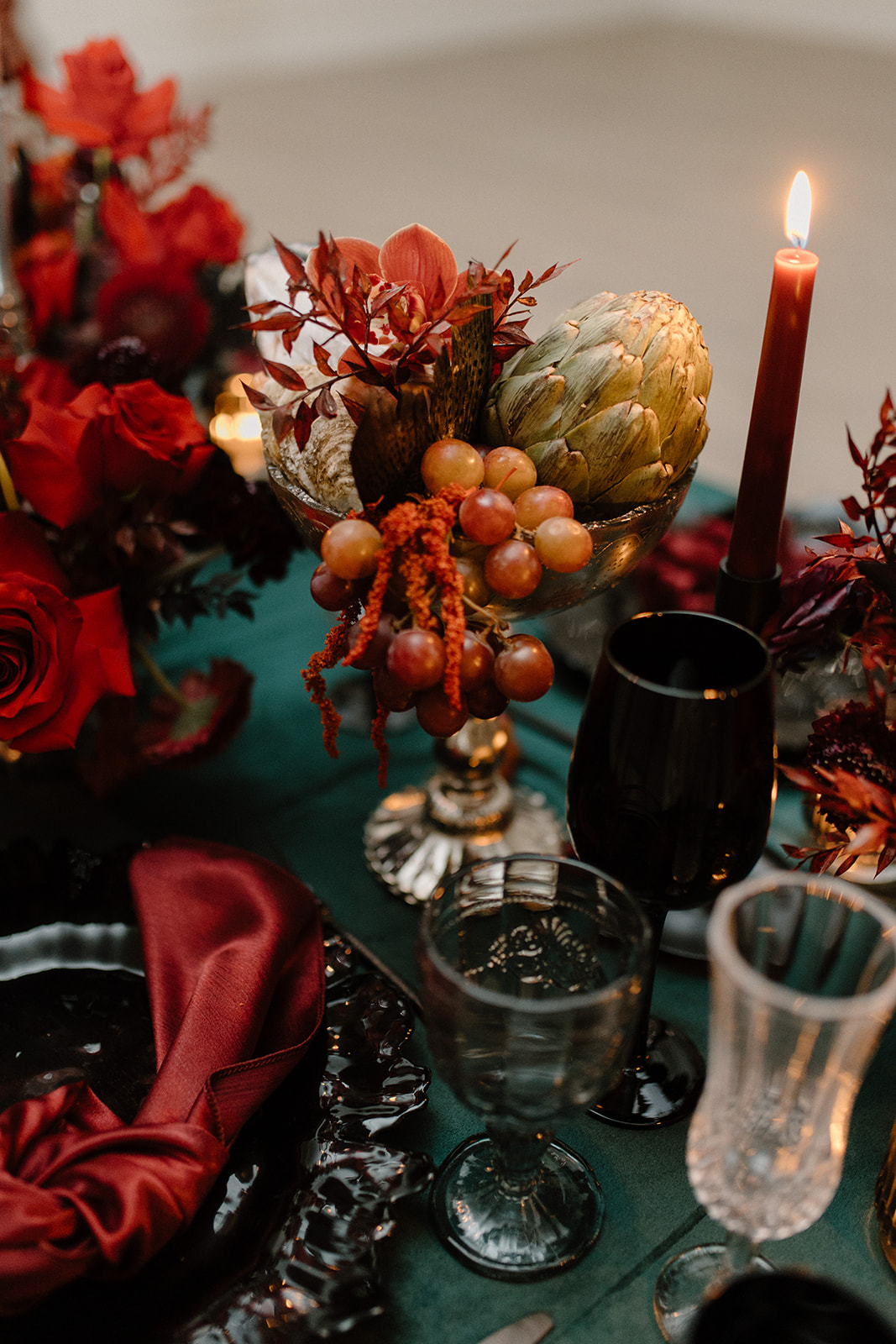 A dimly lit table setting features dark red flowers, an artichoke, and grapes in a vase. A black candle is lit nearby, accompanied by elegant glassware and a red napkin on a dark plate, all on a green tablecloth.