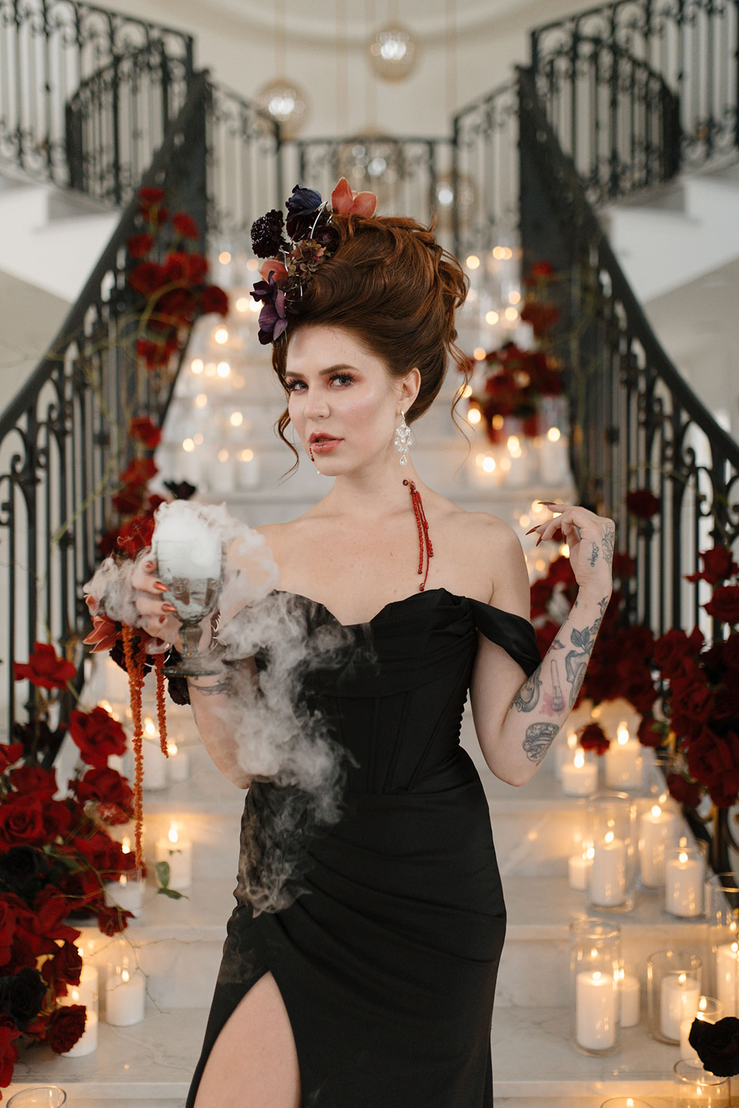 A woman in a black dress stands on a staircase adorned with red roses and lit candles. She holds a smoking chalice and wears a dramatic hairstyle with a floral headpiece. The setting is elegant and theatrical. A Bite of Dark Romance & Gothic Glamour in This Dracula Dinner Party Inspiration