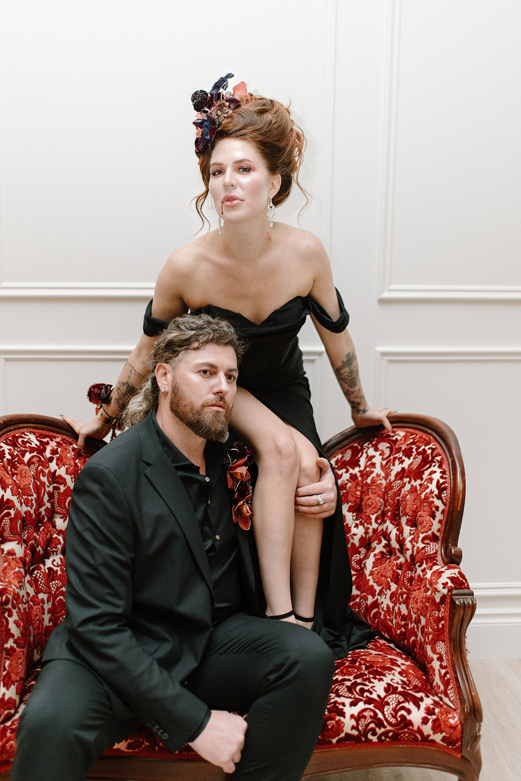 A woman in a black dress and floral headpiece sits on the arm of a red velvet sofa, leaning forward. A man in a black suit with a floral boutonniere sits on the sofa, looking thoughtful. The setting is an elegantly decorated room.