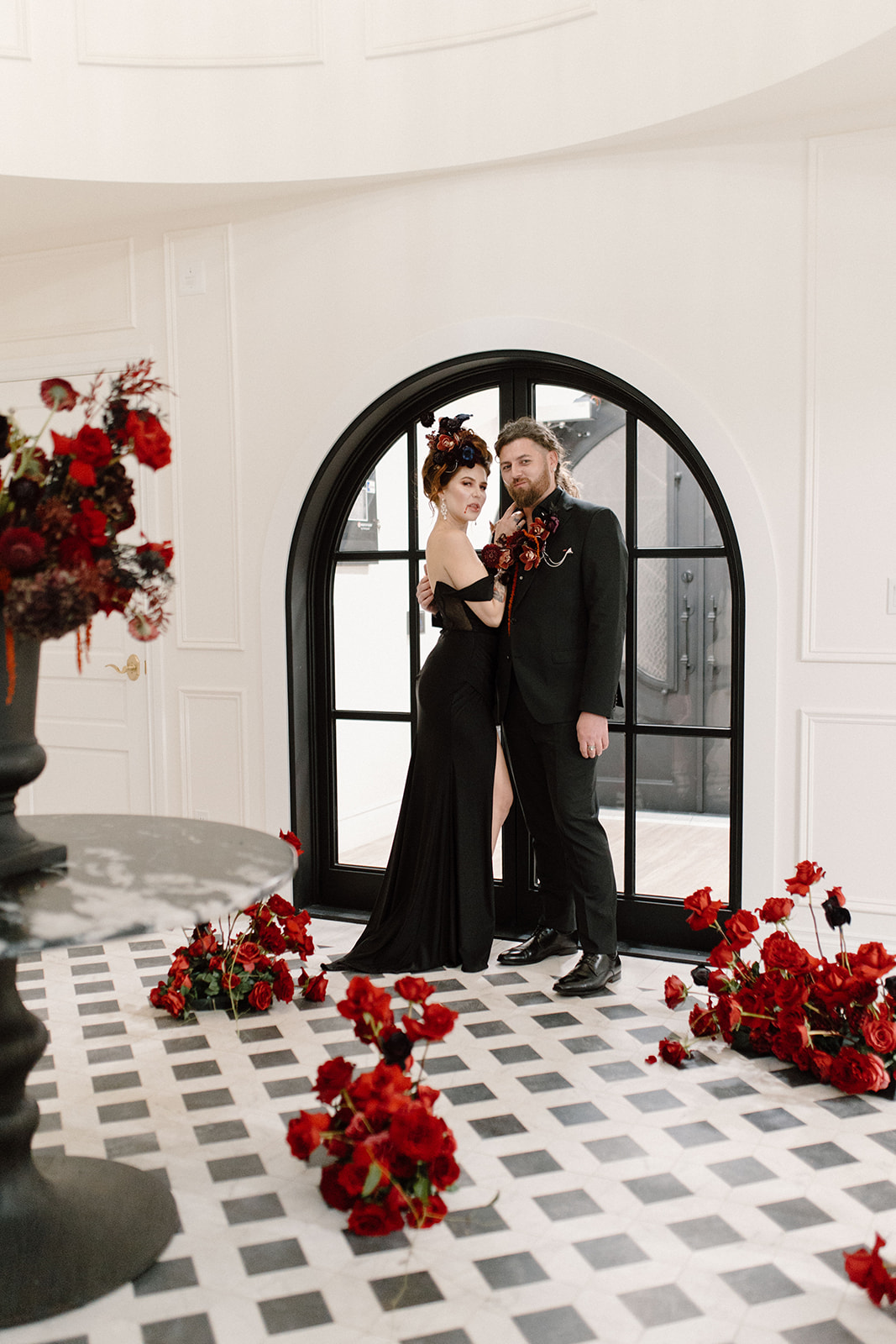 A couple in elegant black attire stands by a large arched window. They are surrounded by red floral arrangements on a checkered floor. The setting is sophisticated, with a modern and romantic atmosphere. A Bite of Dark Romance & Gothic Glamour in This Dracula Dinner Party Inspiration