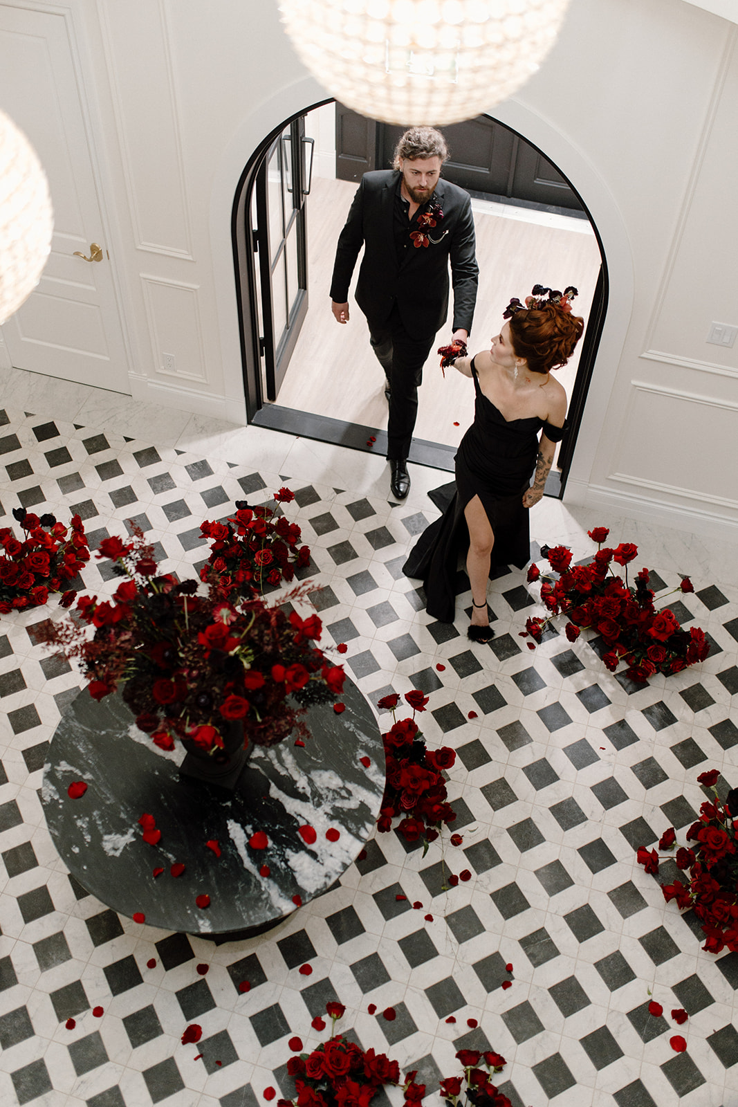 A couple in elegant black attire walks across a checkered floor adorned with scattered red rose petals. A round table with a tall arrangement of red roses enhances the romantic ambiance of the room.