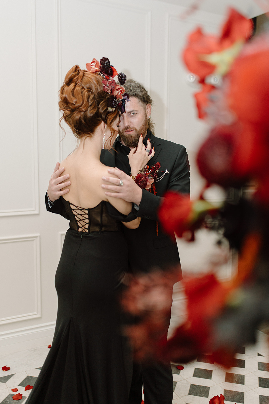 A couple dressed elegantly in black, embracing in a warmly decorated setting. The woman has a floral headpiece, and rose petals are scattered on the floor and foreground. The atmosphere is romantic and sophisticated.