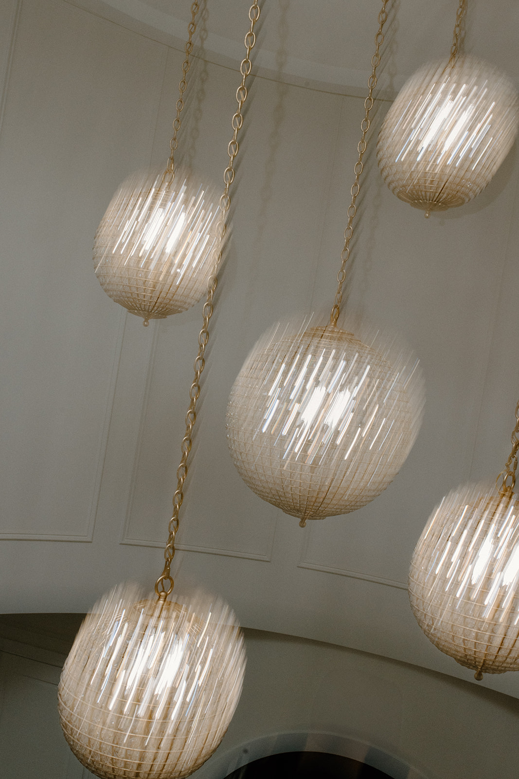 A set of five spherical pendant lights with gold chains hang from a ceiling. The lights have a textured, translucent design that emits a soft, warm glow, casting delicate shadows on the white background.