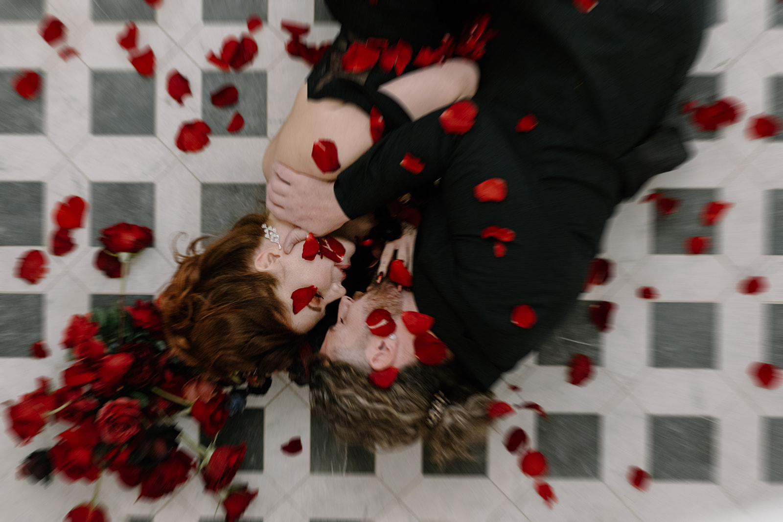 A couple lies on a checkered floor, surrounded by scattered red rose petals. They are embracing closely, with red roses nearby, creating a romantic and intimate atmosphere.