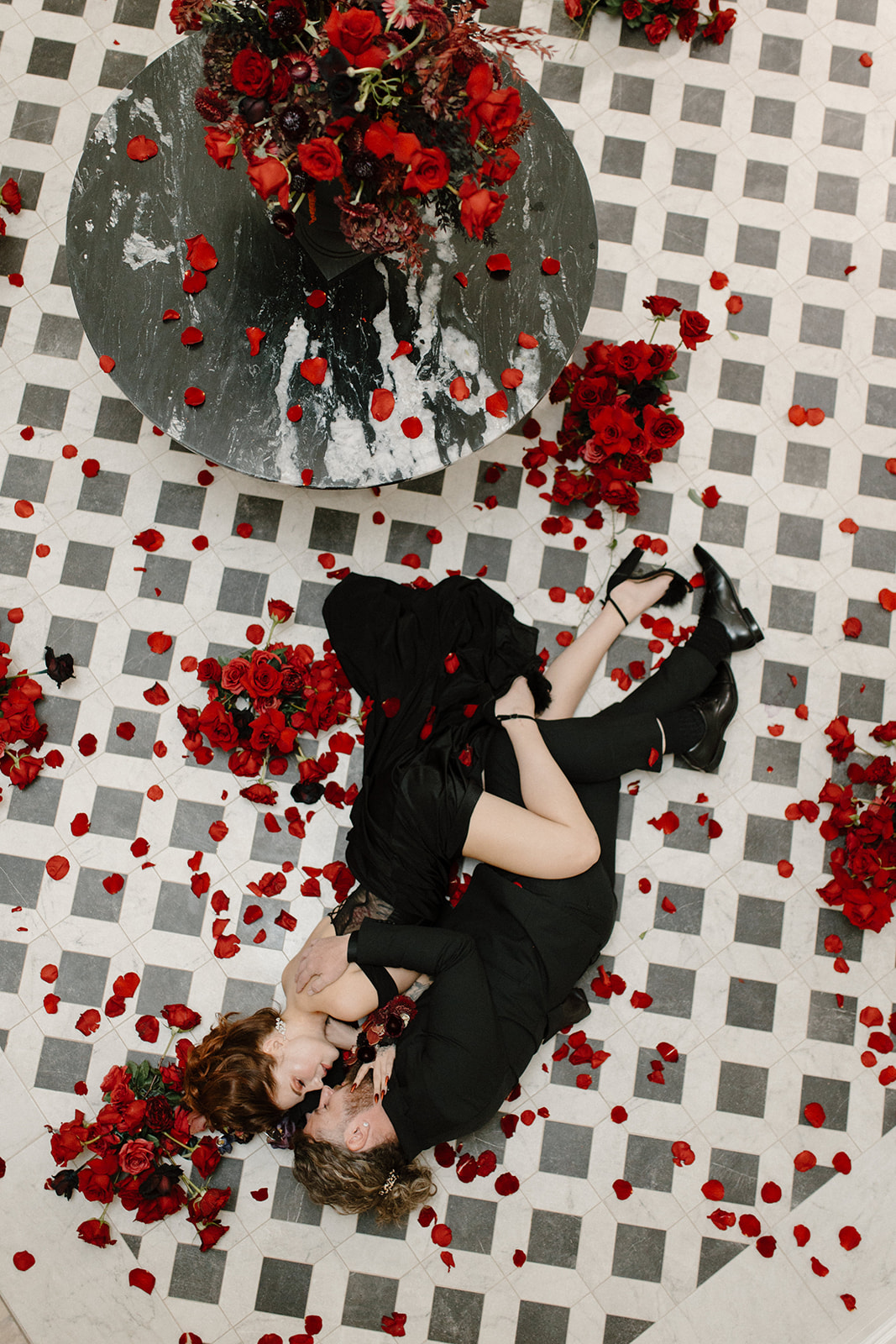 A couple lies on a checkered floor surrounded by scattered red rose petals. They embrace lovingly, dressed in black, with more roses on a round black table above them. The scene is romantic and artistic.