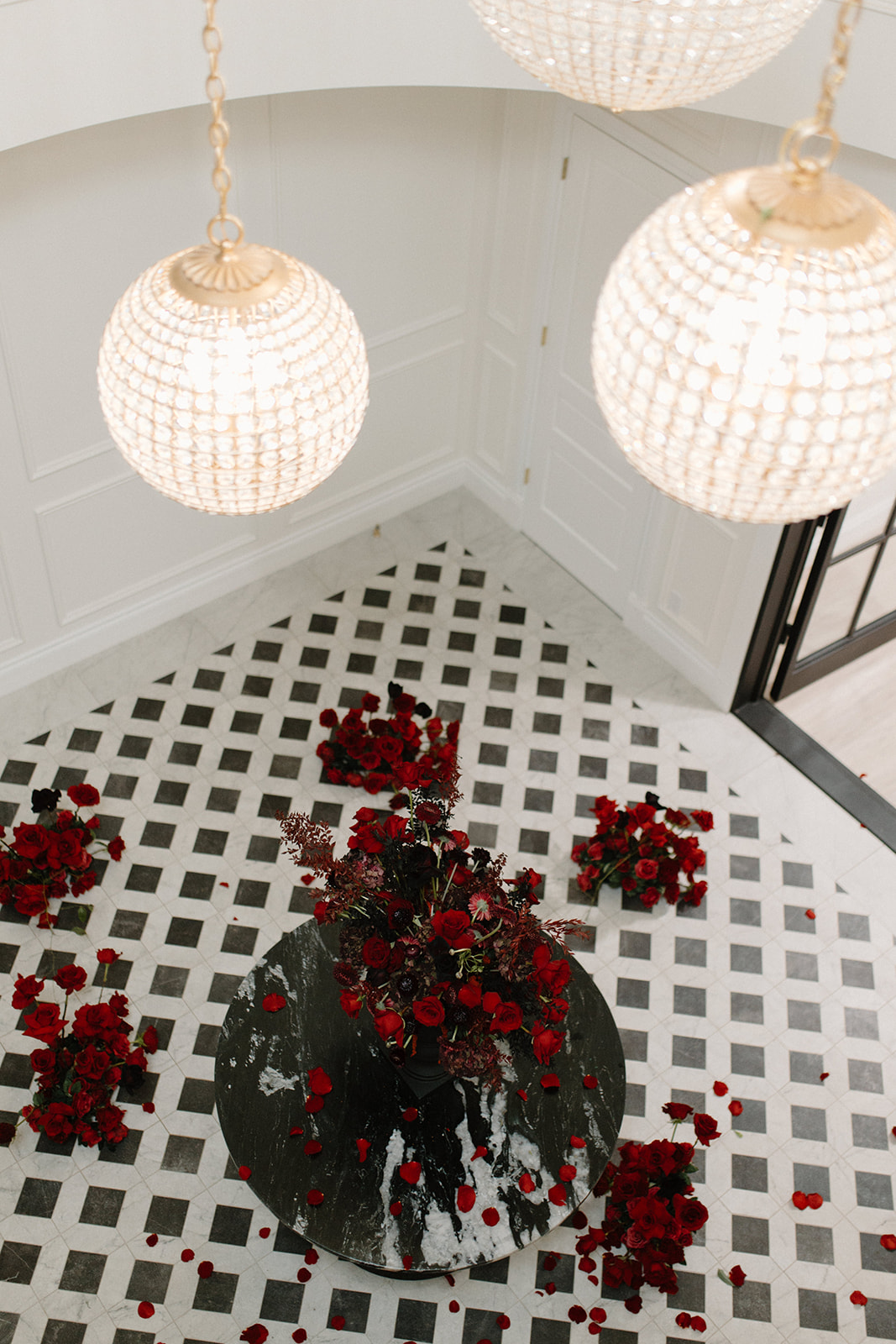 A top-down view of a floor with a black and white checkered pattern. Red roses are arranged in the center and scattered around. Two ornate chandeliers hang above, casting light on the scene. The walls are white, creating a bright and elegant atmosphere.