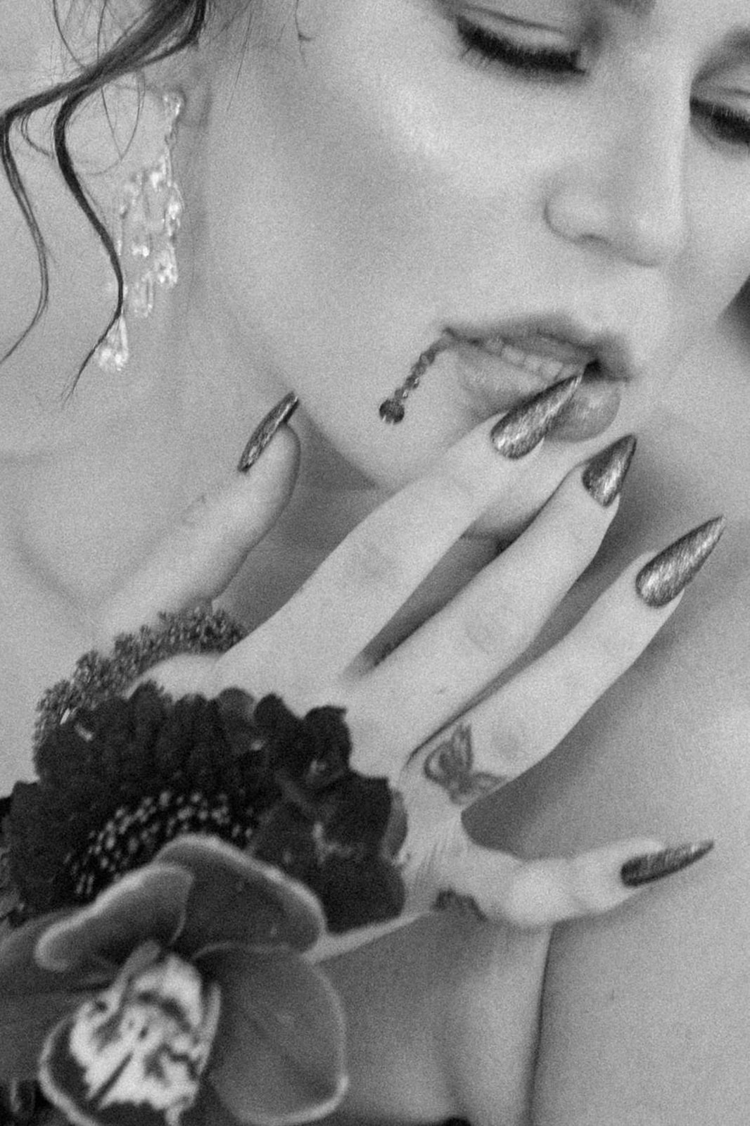 A black and white photo of a woman with long, dark nails touching her lip and chin, where liquid trickles. She has floral and butterfly tattoos on her hand, and there's a large flower embellishment on her wrist. She wears a dangly earring.