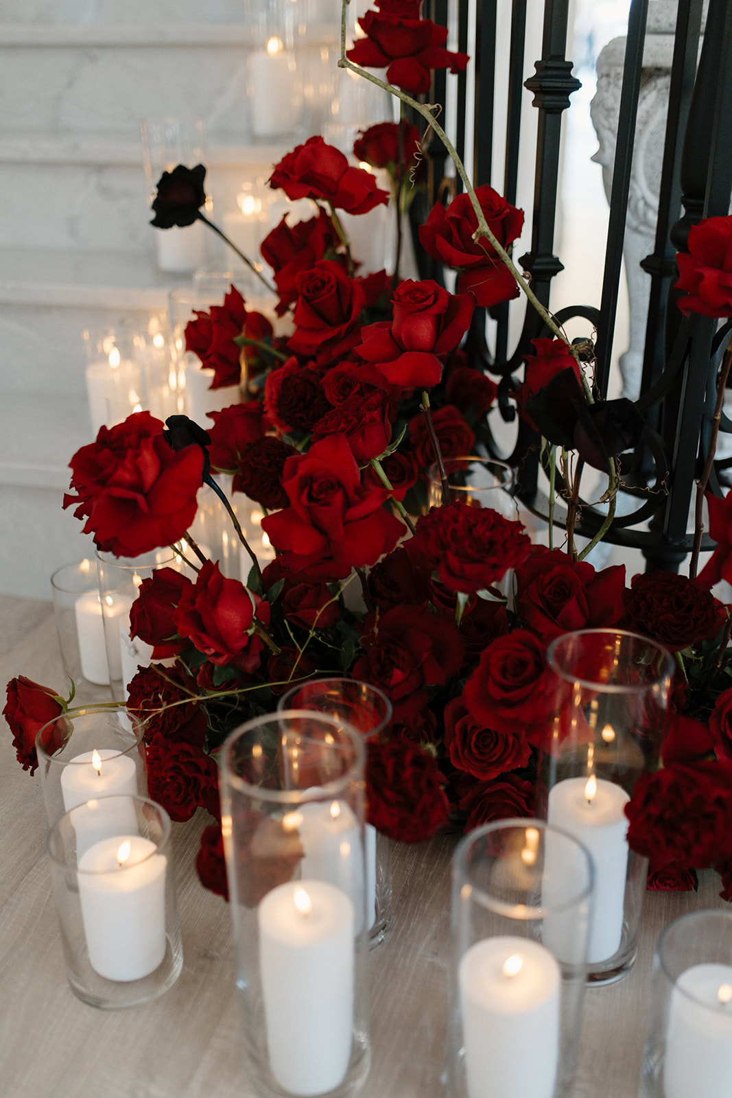 A luxurious arrangement of red roses is displayed in front of a staircase, surrounded by numerous lit white candles in glass holders. The scene is elegantly set, suggesting a romantic or sophisticated setting.