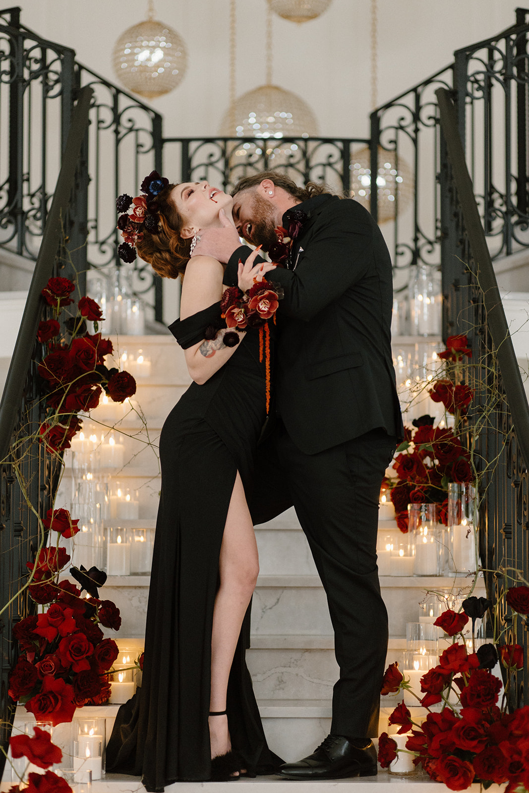 A couple dressed in formal black attire poses playfully on a staircase adorned with red roses and lit candles. TA Bite of Dark Romance & Gothic Glamour in This Dracula Dinner Party Inspiration