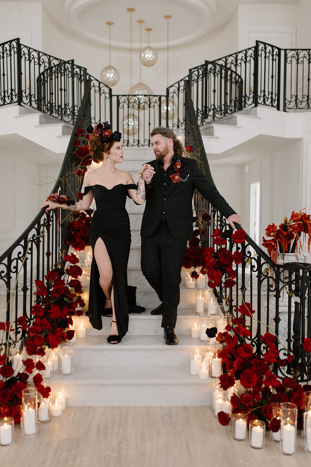 A couple in elegant black attire descends a grand staircase adorned with red roses and candles. The woman wears a dress with a slit and a floral headpiece, while the man sports a matching suit. The setting is sophisticated and romantic. A Bite of Dark Romance & Gothic Glamour in This Dracula Dinner Party Inspiration