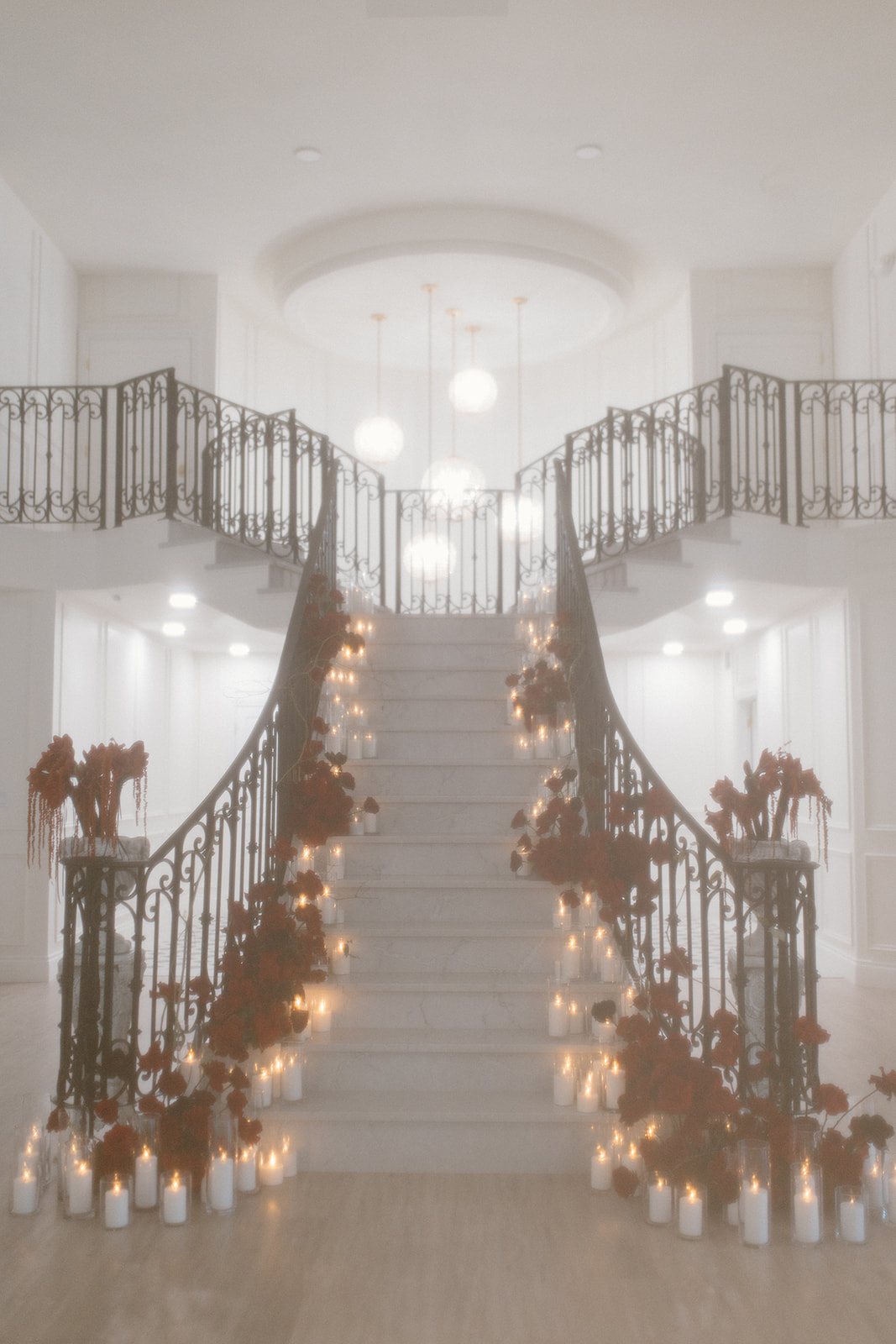 A grand staircase with elegant iron railings adorned with red flowers and lit candles, creating a romantic atmosphere. Hanging lights illuminate the space, adding to the ethereal ambiance.