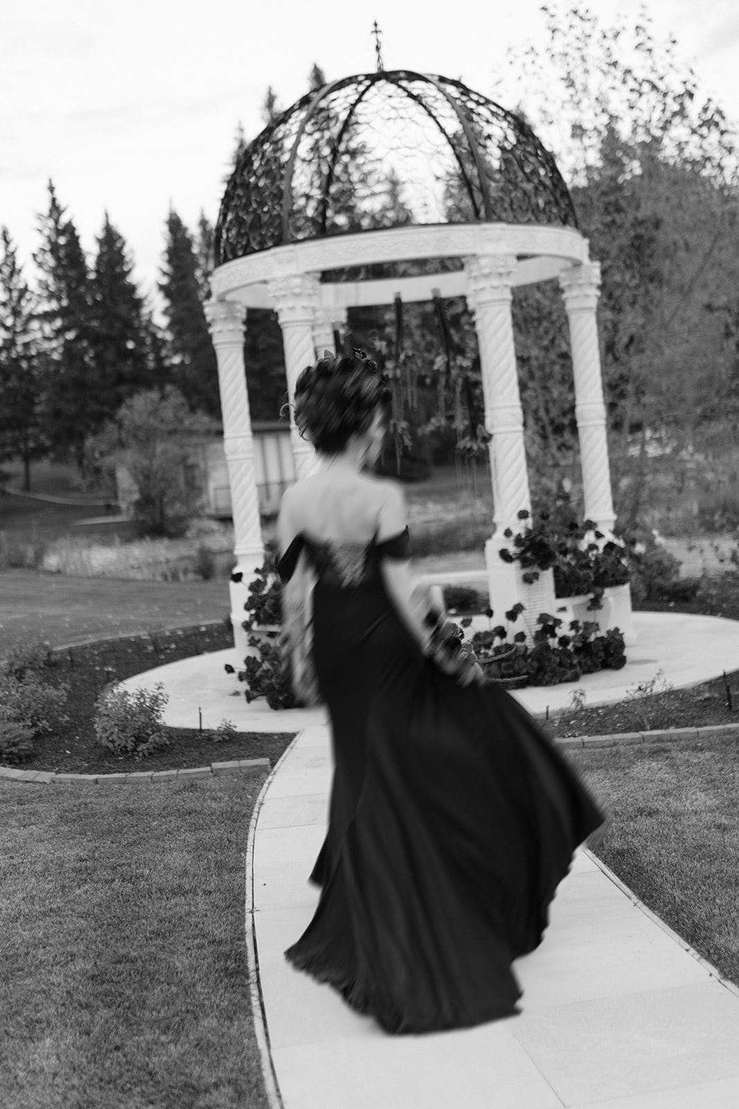 A woman in a flowing black gown walks along a path toward a decorative garden gazebo. The image is in black and white, with trees and manicured landscaping in the background. Halloween inspiration