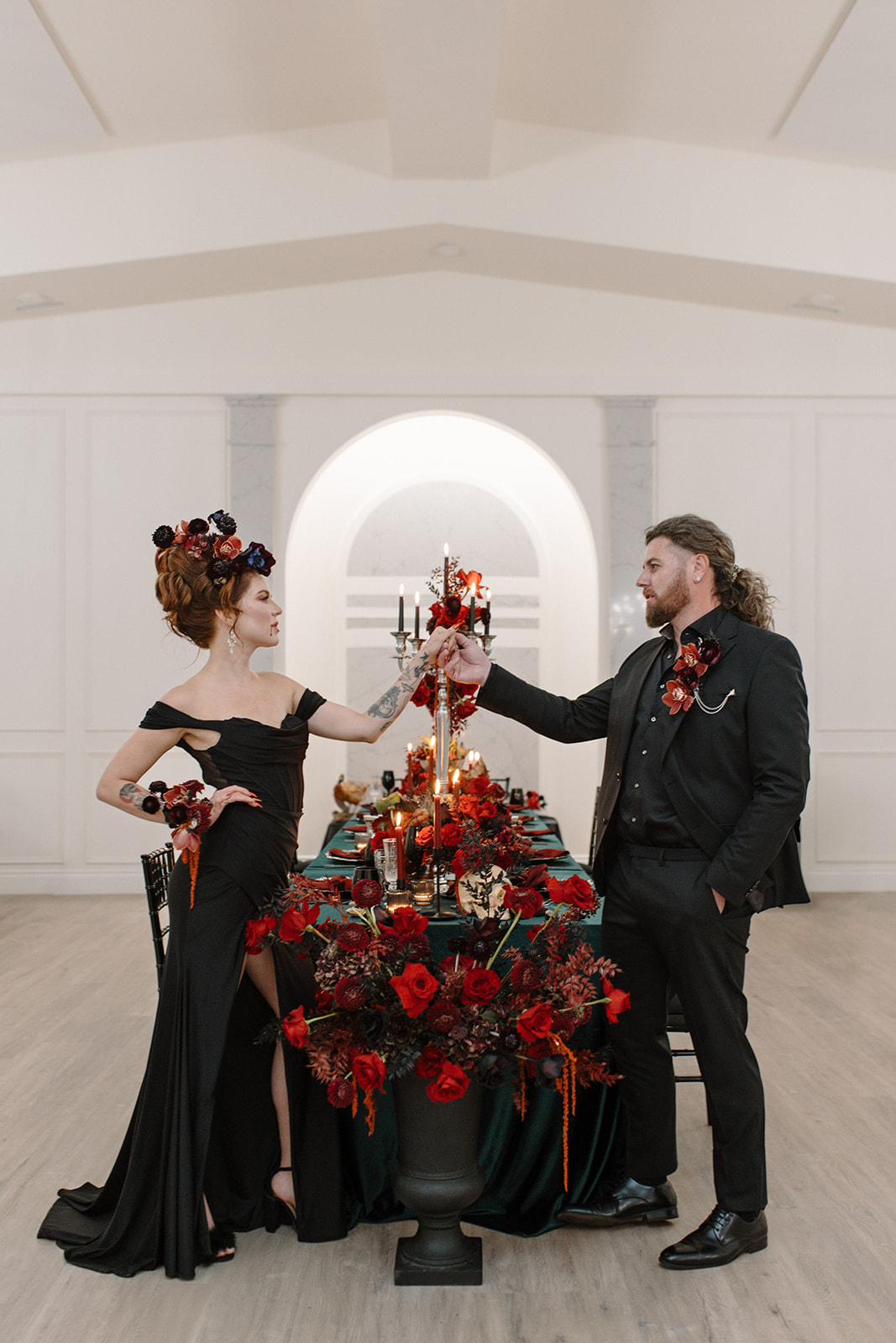 A couple dressed in black attire clinks glasses at a lavishly decorated table with red and black flowers, candles, and elegant tableware in a bright, white room with an arched niche in the background. A Bite of Dark Romance & Gothic Glamour in This Dracula Dinner Party Inspiration