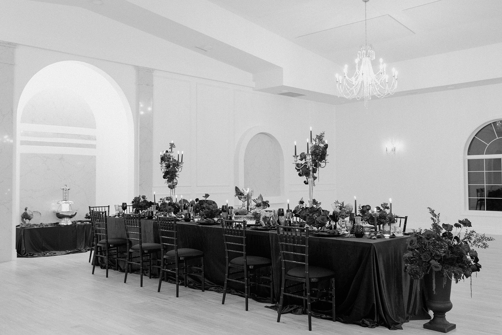 A black and white photo of an elegant dining setup. A long table with dark tablecloth and ornate centerpieces is surrounded by chairs. The room features high ceilings, a chandelier, and arched windows, creating a sophisticated atmosphere.