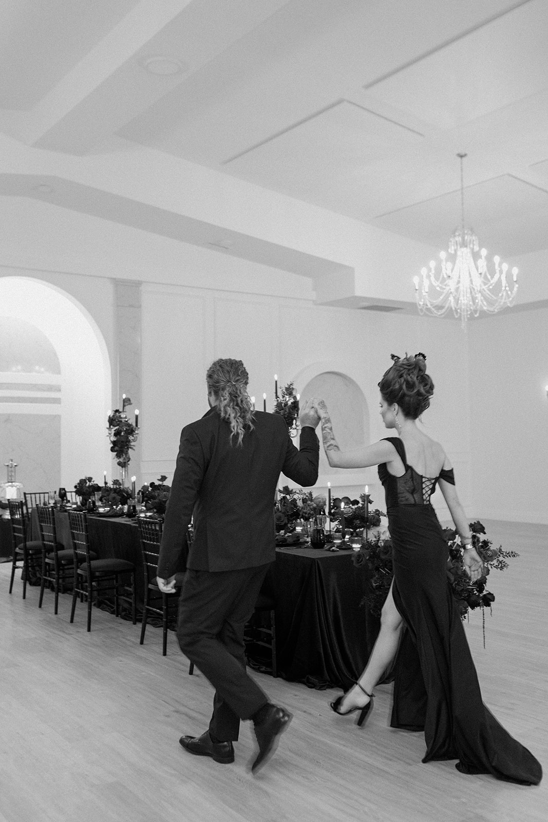 A couple dressed in formal attire walks hand in hand through an elegantly decorated banquet hall. The room features a long dining table with black tablecloths, candle arrangements, and a chandelier hanging from the ceiling. The image is in black and white. A Bite of Dark Romance & Gothic Glamour in This Dracula Dinner Party Inspiration