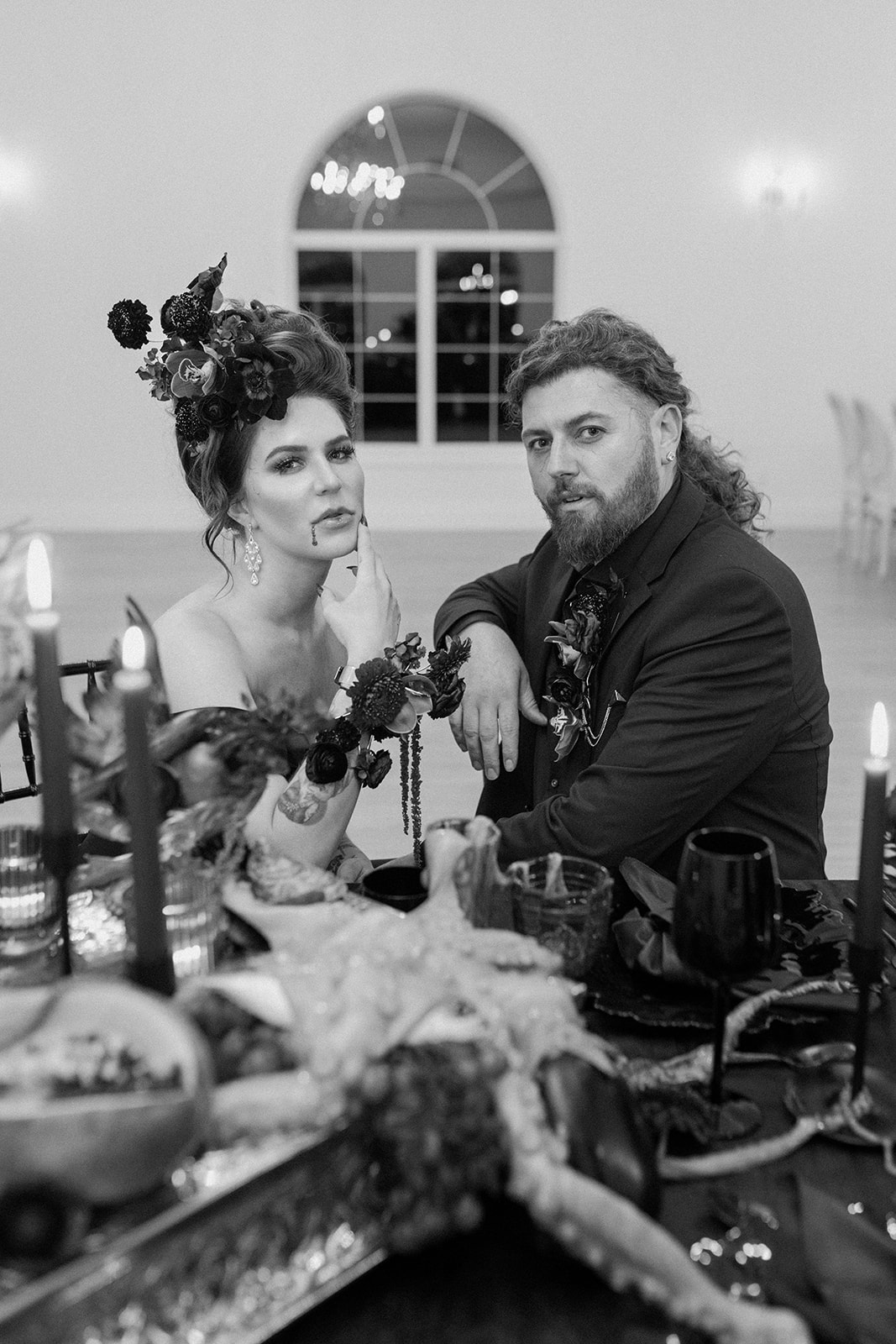 A Bite of Dark Romance & Gothic Glamour in This Dracula Dinner Party Inspiration. A couple sits at a dimly lit, elegantly decorated table. The woman wears a floral headpiece and dark attire, while the man has a similar style. They both have a serious expression, surrounded by candles and ornate tableware.