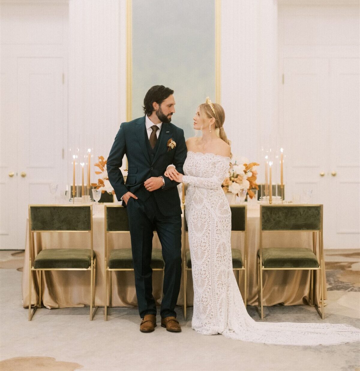 A couple stands in a lavish, elegantly decorated room. The groom wears a dark suit, and the bride wears a lace off-the-shoulder gown. Behind them is a table adorned with flowers and candles, set against a backdrop of ornate white and gold decor.