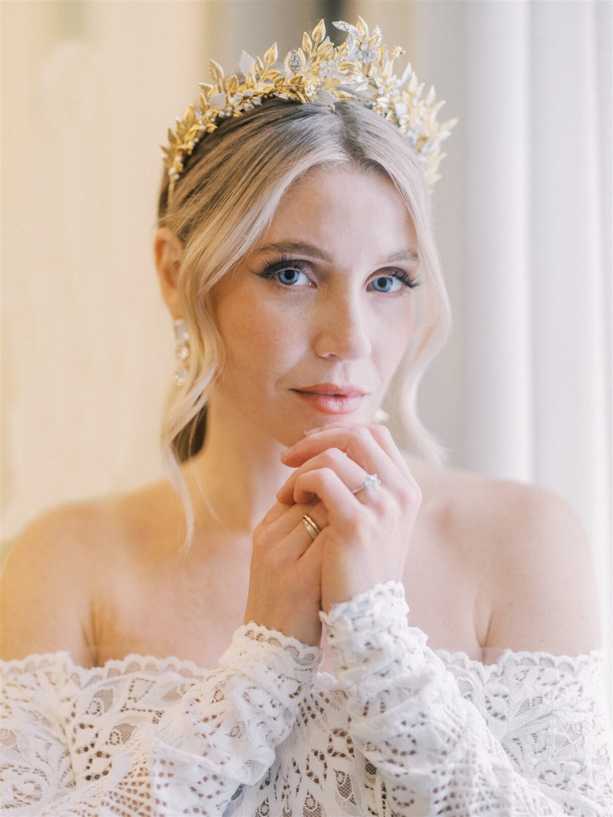 A woman wearing a lace off-the-shoulder dress and a golden leaf crown looks softly at the camera, with her hands gently clasped under her chin. Her hair is styled in loose waves, and she is standing in a softly lit room.