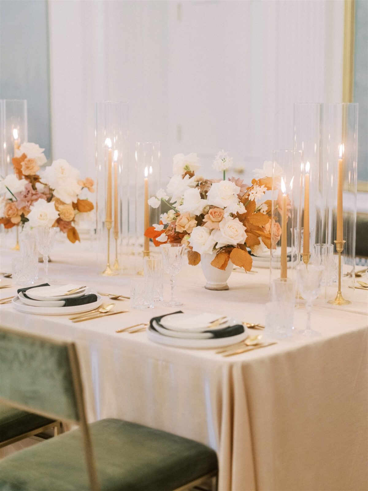 Elegant dining table setup with white and gold dinnerware, surrounded by tall, thin candles in glass holders. Floral centerpieces with white and blush roses and autumn leaves add a touch of color. The overall ambiance is sophisticated and welcoming.