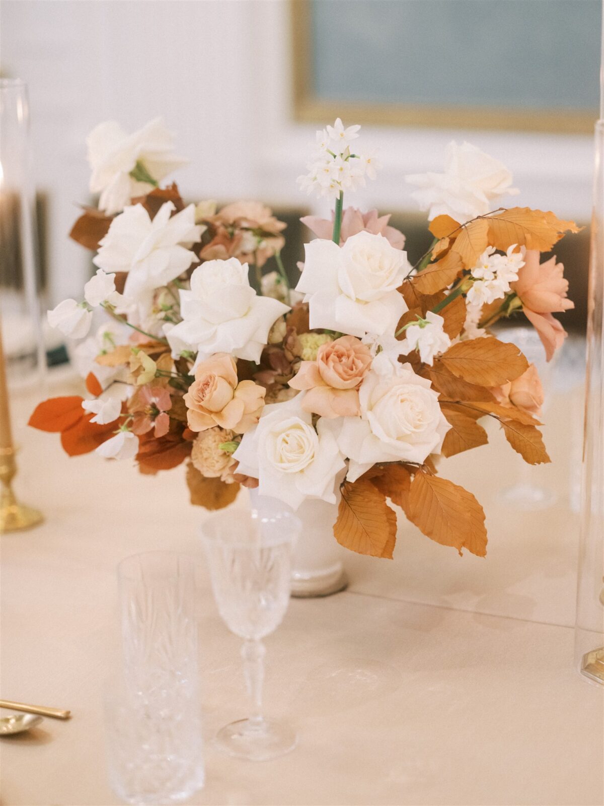 A floral arrangement with white and peach roses, surrounded by autumn leaves in warm tones. It sits on a softly lit table with crystal glasses and a gold candle holder, creating an elegant and cozy atmosphere.