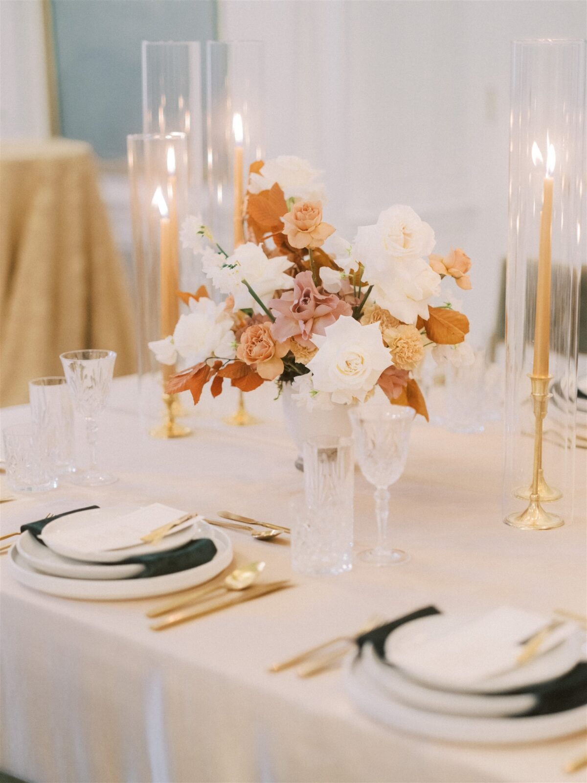 Elegant table setting with tall candles, crystal glasses, and fine china. A centerpiece features a bouquet of white and peach flowers. The ambiance is sophisticated and inviting.