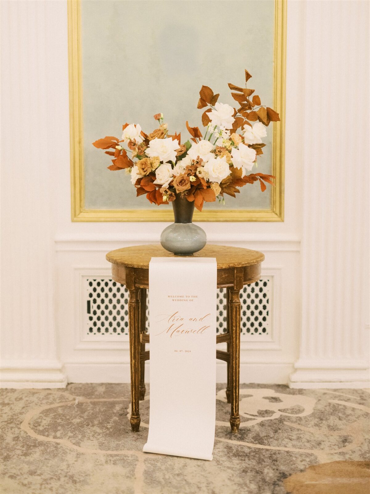 A floral arrangement of white, toffee, and burnt orange flowers in a vase sits on a small round wooden table. A long white cloth is the welcome sign for the wedding, with calligraphy hangs from the table, reading, "Alex and Meredith," with a date below. The backdrop is an elegant room with a muted color scheme.