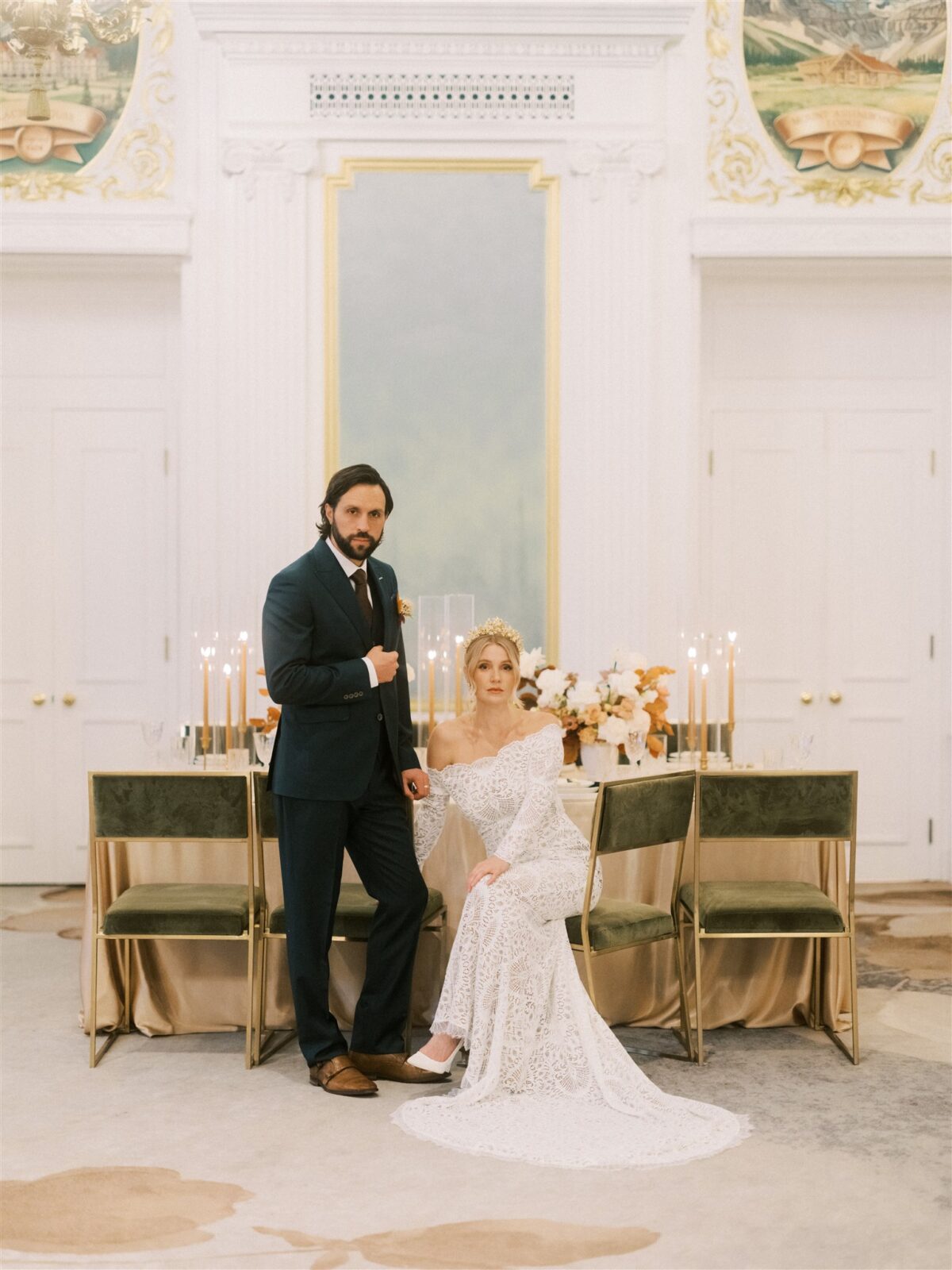 A couple poses elegantly indoors near a table adorned with floral arrangements and candles. The woman is seated, wearing a white, off-the-shoulder lace gown and a crown. The man stands beside her in a dark suit. The setting has a luxurious, classic decor.