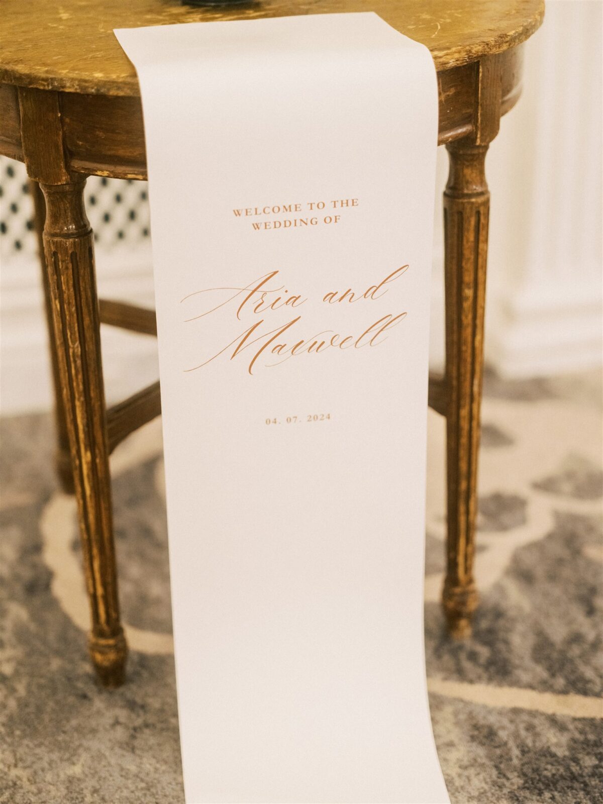 A wedding sign on a table reads "Welcome to the wedding of Aria and Maxwell" with the date "04.07.2024" in elegant script. The sign is placed on a rustic wooden table with a patterned carpet underneath.