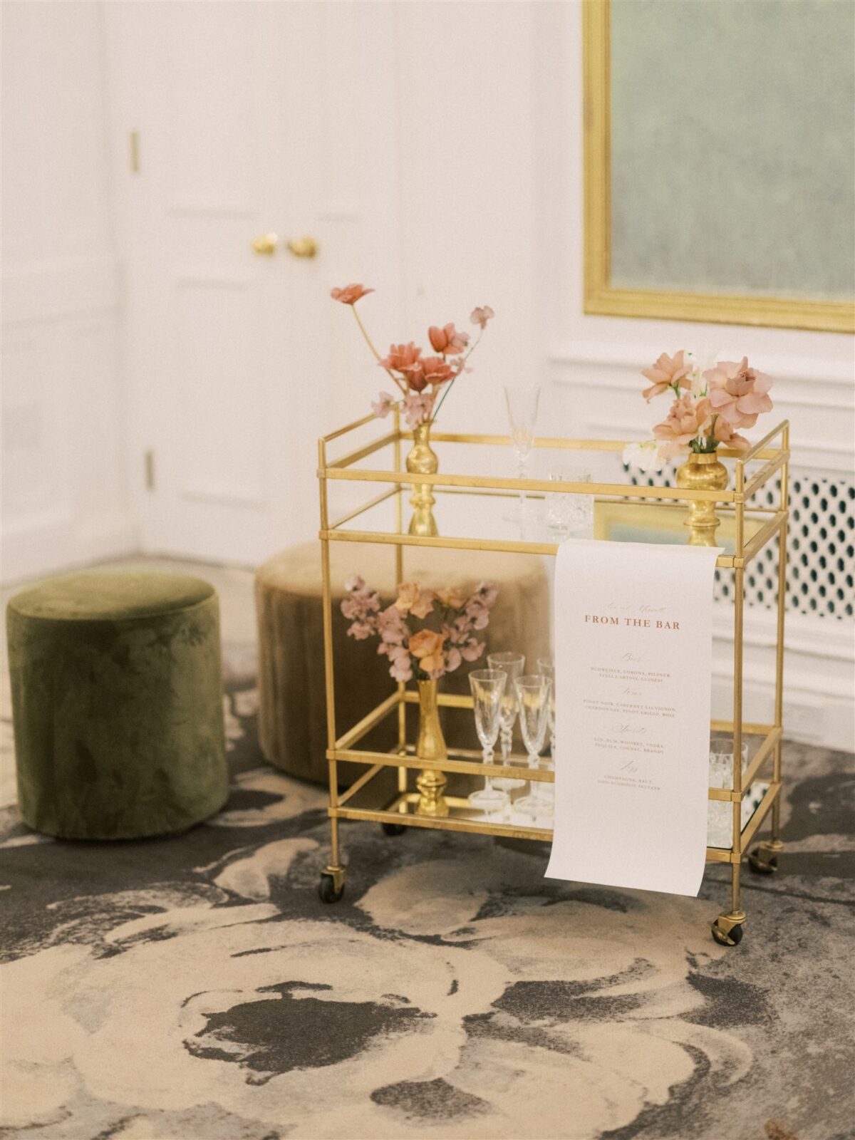 A stylish gold bar cart with flowers in vases and empty glasses on a floral-patterned rug in an elegant room. A menu hangs from the cart. Green and beige ottomans are nearby, and a white wall with a large framed artwork is in the background.
