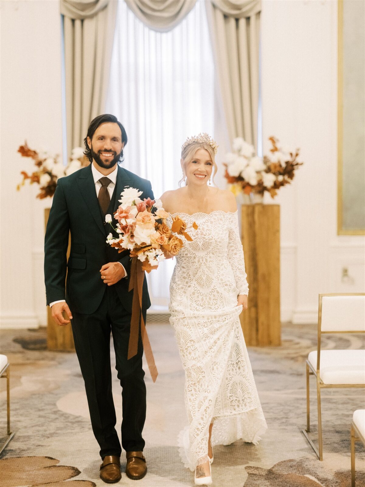 A bride and groom joyfully walk down the aisle in an elegantly decorated room. The bride wears a lace gown and holds a bouquet of autumn flowers, while the groom is in a dark suit. The background features white flowers and wood accents.