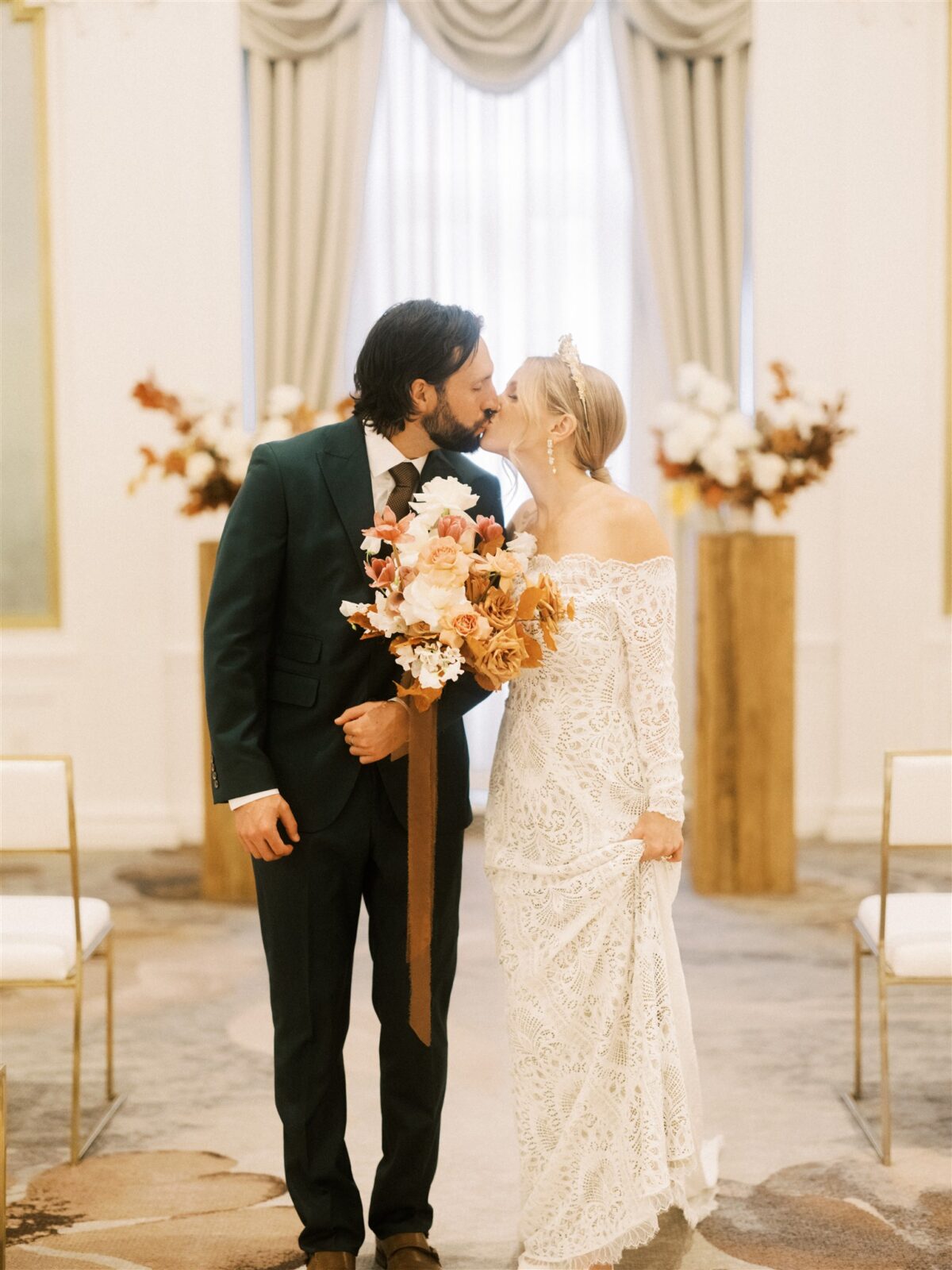A couple in wedding attire shares a kiss in an elegant room. The bride holds a bouquet of white and orange flowers, wearing a long lace gown. The groom is in a dark suit. Floral arrangements and chairs line the aisle behind them.
