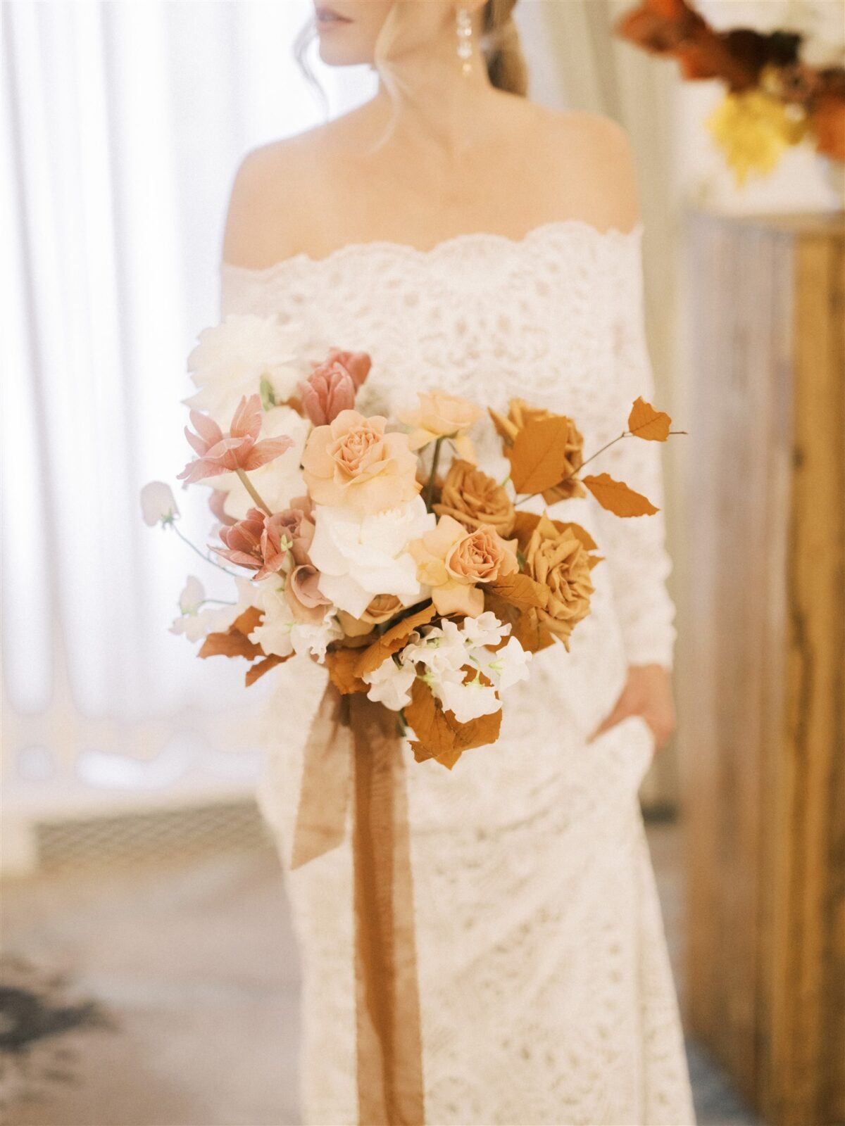A bride in an off-the-shoulder lace gown holds a bouquet of roses and other flowers in soft pink, peach, and rust hues. She stands indoors, with light streaming in from a window behind her.
