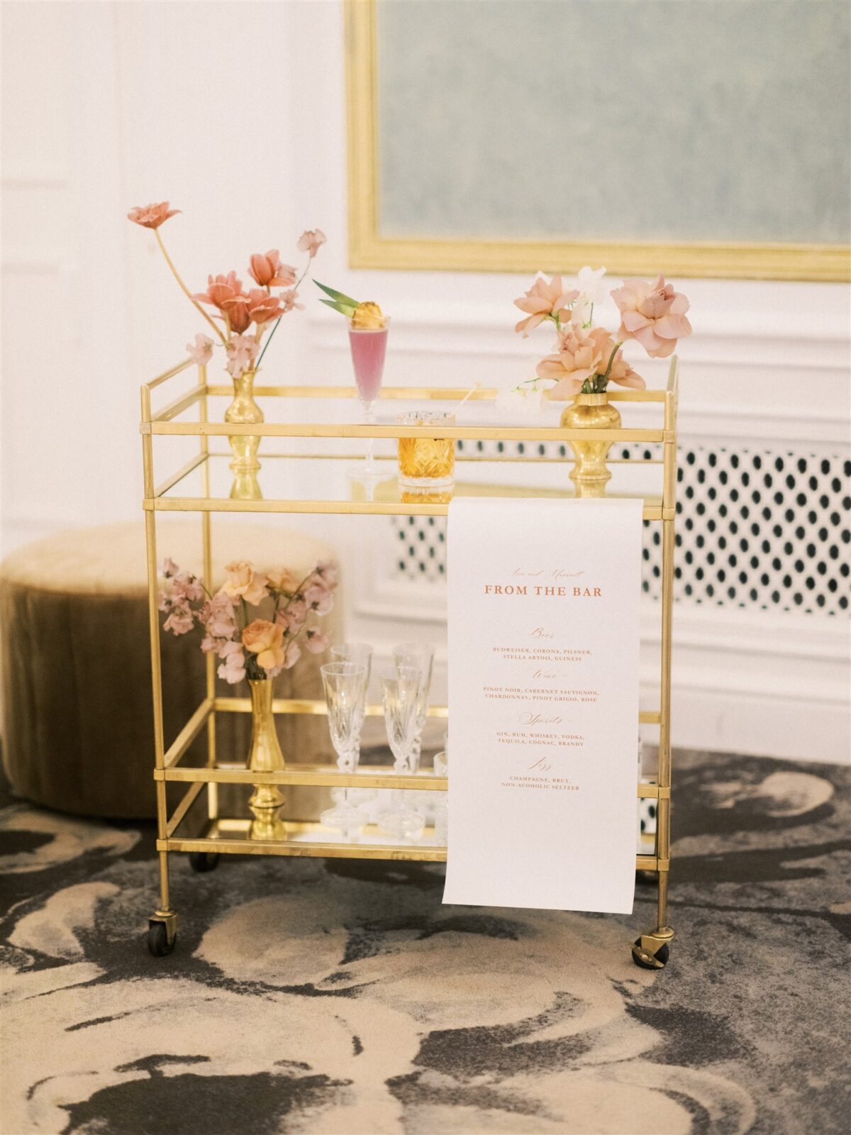 A stylish gold bar cart with flowers in vases and empty glasses on a floral-patterned rug in an elegant room. A bar menu hangs from the cart. Green and beige ottomans are nearby, and a white wall with a large framed artwork is in the background.