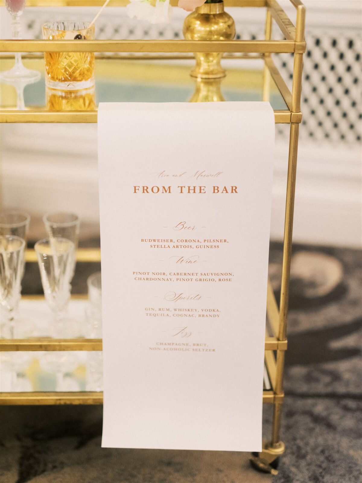 A stylish gold bar cart with flowers in vases and empty glasses on a floral-patterned rug in an elegant room. A bar menu hangs from the cart. Green and beige ottomans are nearby, and a white wall with a large framed artwork is in the background.
