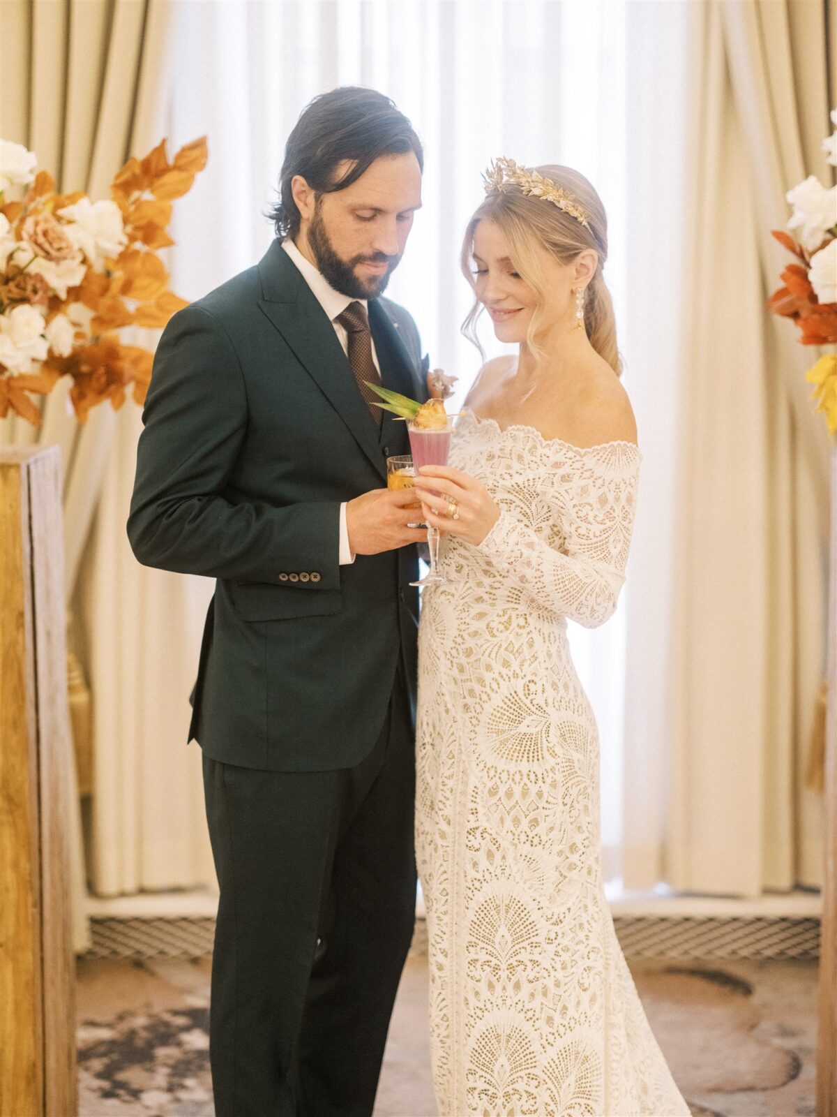 In a warmly lit room, a couple embodies elegant Fairmont wedding inspiration. The groom dons a dark green suit, and the bride shines in a lace gown with a floral headpiece. They toast with colorful cocktails amidst lush floral arrangements.