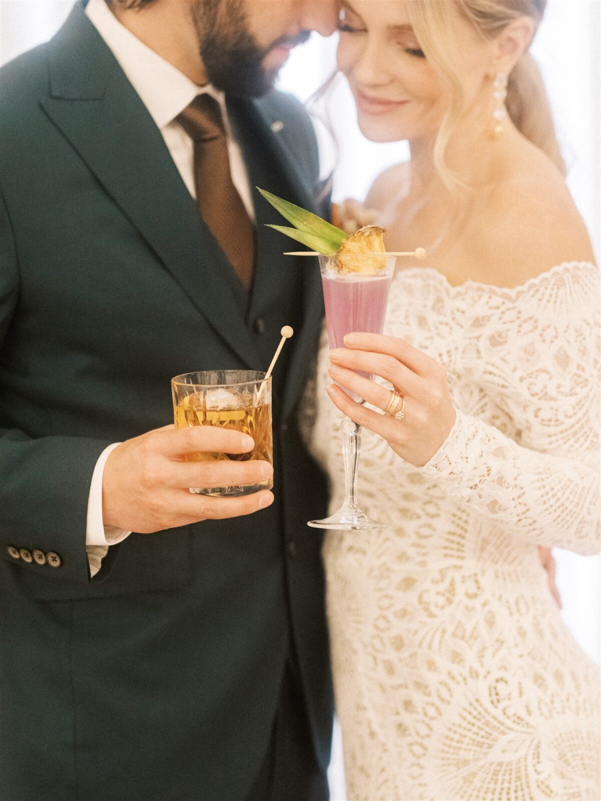In a warmly lit room, a couple embodies elegant Fairmont wedding inspiration. The groom dons a dark green suit, and the bride shines in a lace gown with a floral headpiece. They toast with colorful cocktails amidst lush floral arrangements.