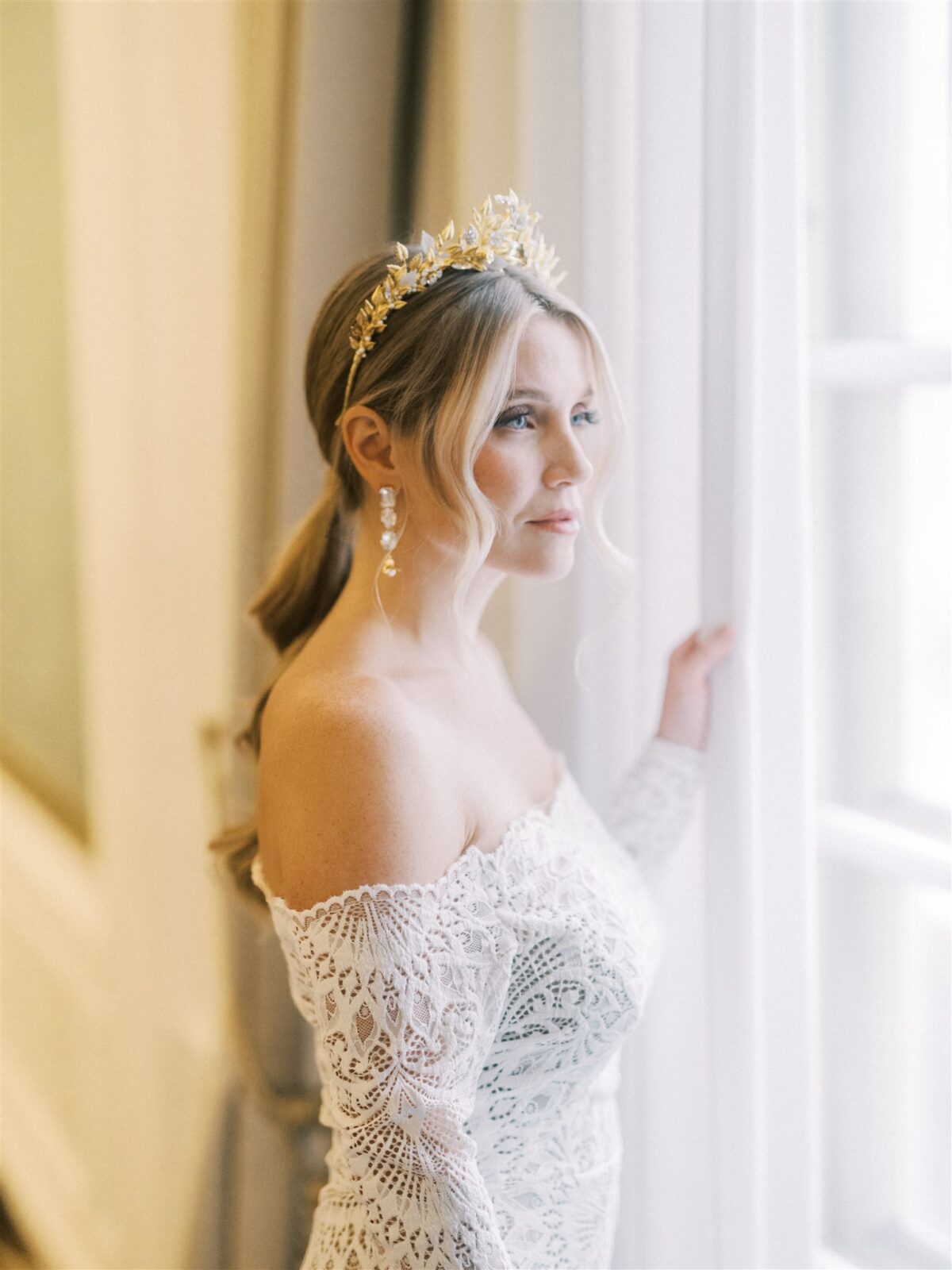A bride in an off-the-shoulder, lace wedding dress stands by a window. She wears a gold floral crown and dangling earrings, with her hair styled in a loose ponytail. Soft natural light illuminates the scene.