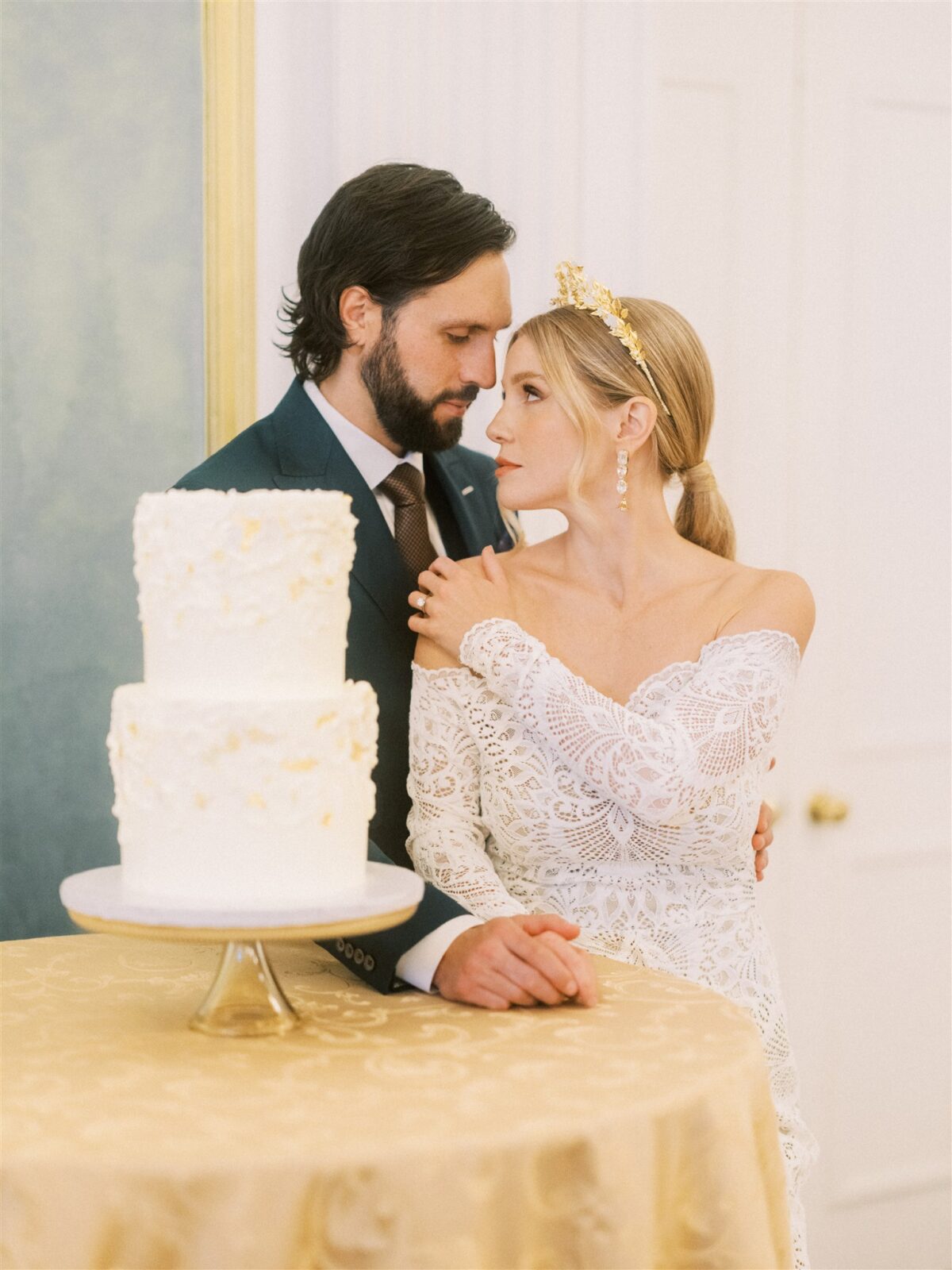 A bride and groom stand closely together in an elegant setting, with the bride wearing a lace off-shoulder gown and a golden tiara. A two-tier white cake is on a table in front of them.