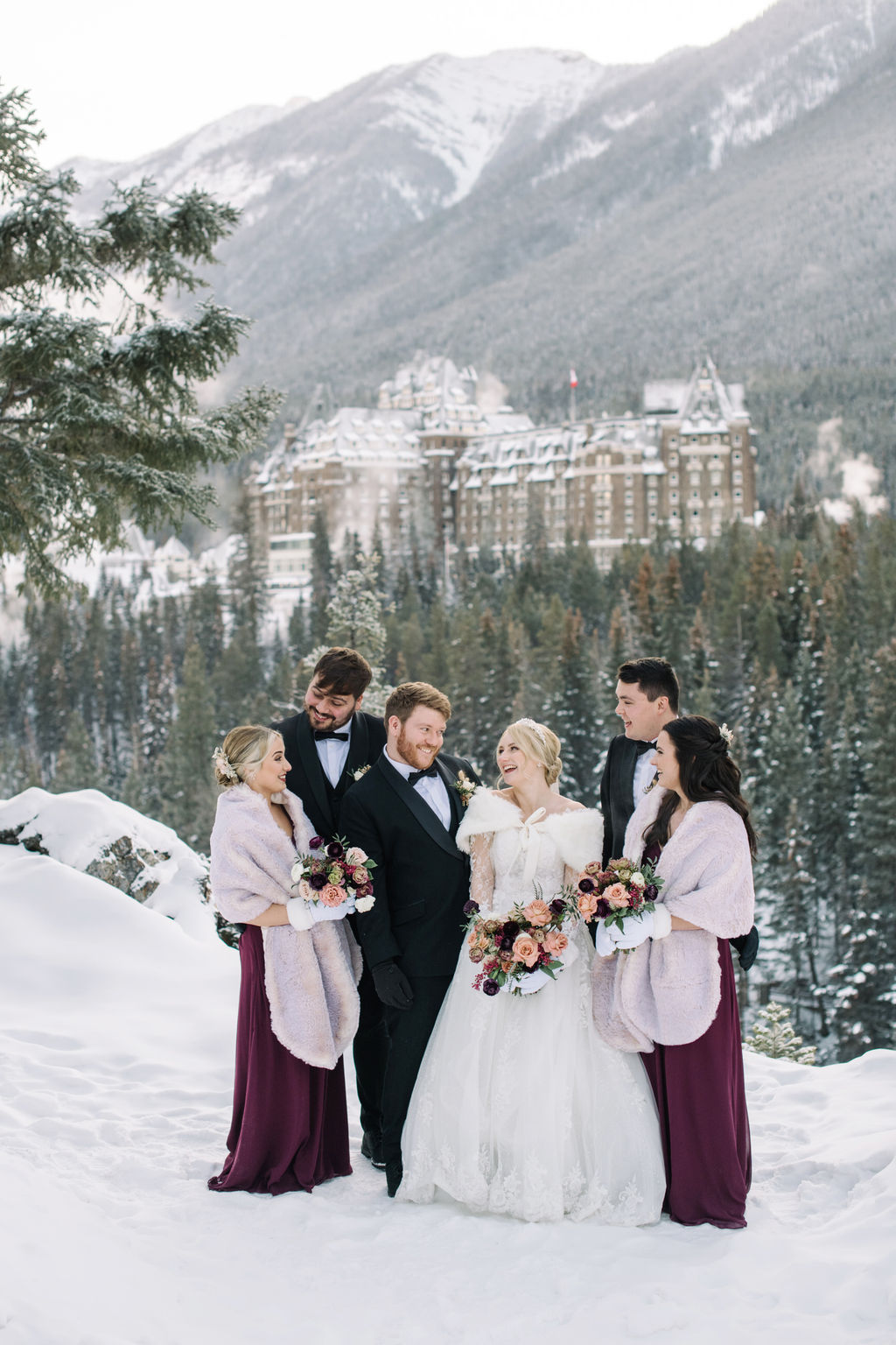 Wedding party poses in the snowy mountains with Fairmont Banff Springs behind them.