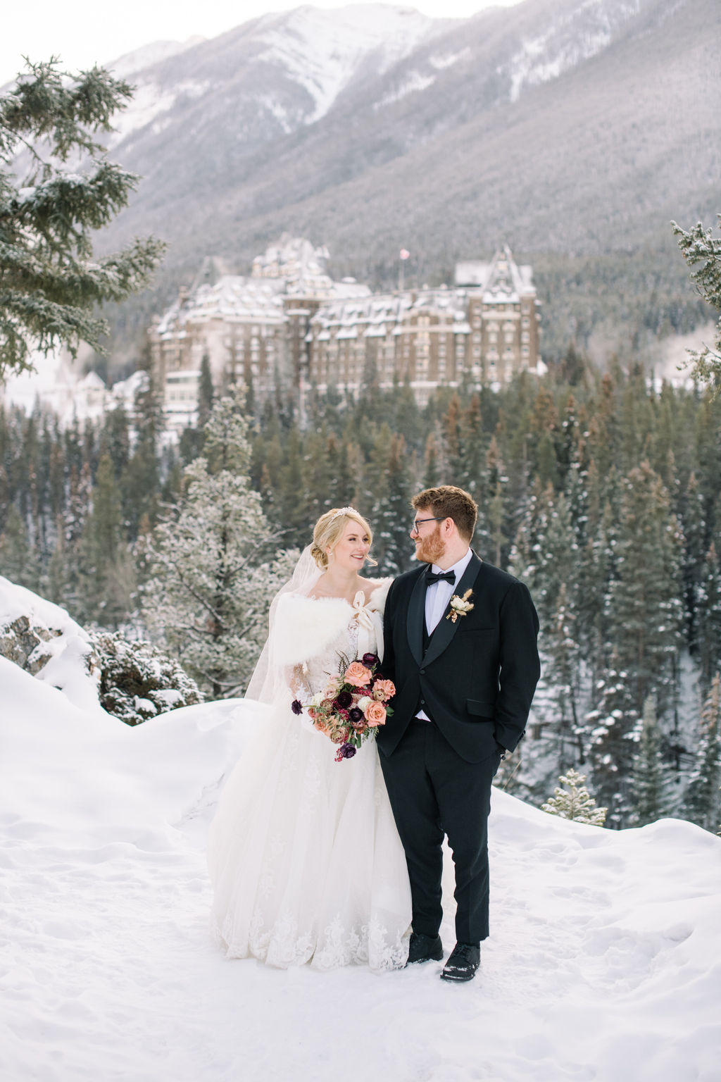 Bride and groom pose in the snow-dusted mountains with Fairmont Banff Springs in the distance, capturing the magic of a winter wedding in Banff.