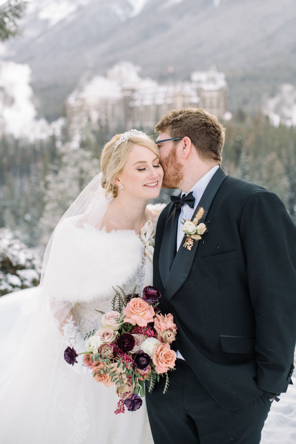 Bride and groom pose in the snow-dusted mountains with Fairmont Banff Springs in the distance, capturing the magic of a winter wedding in Banff.